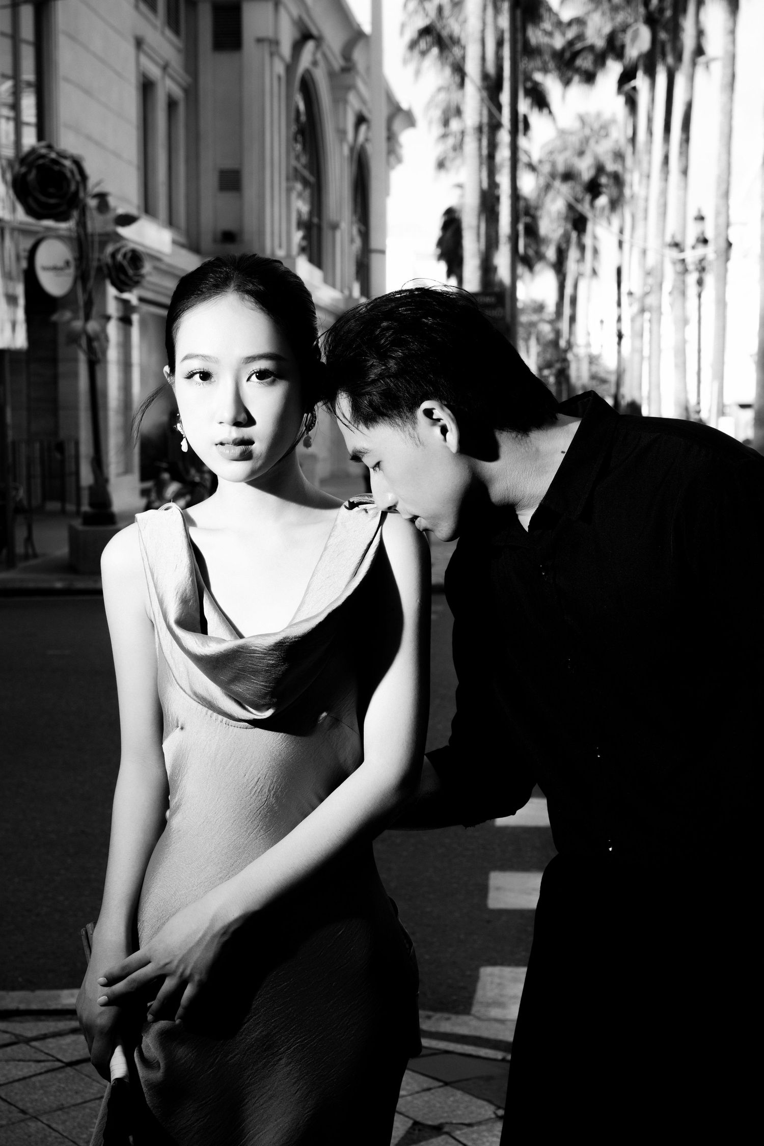 Black and white photo of an elegant Asian couple posing on a city street, woman in satin dress, man in black suit.