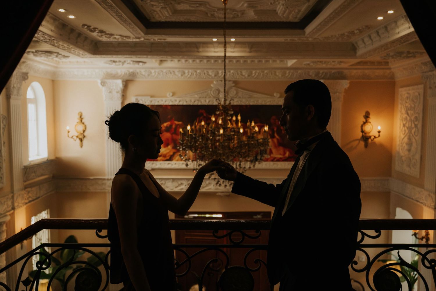 Silhouetted couple holding hands on elegant ballroom balcony with ornate chandelier and warm golden lighting.