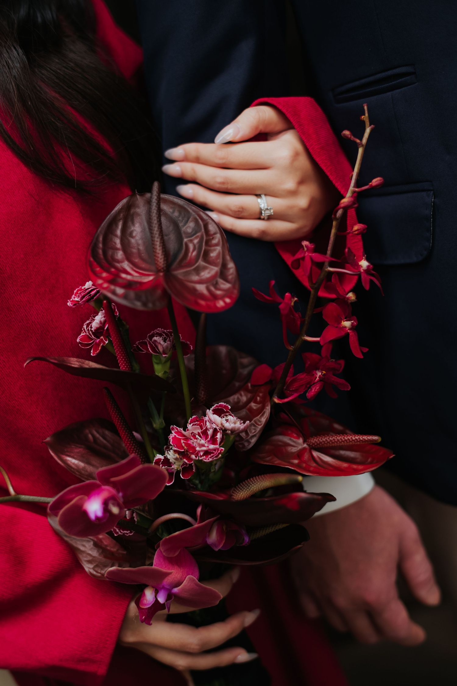 Couple holding a dark red bouquet with anthuriums and orchids, woman wearing an engagement ring in red coat.