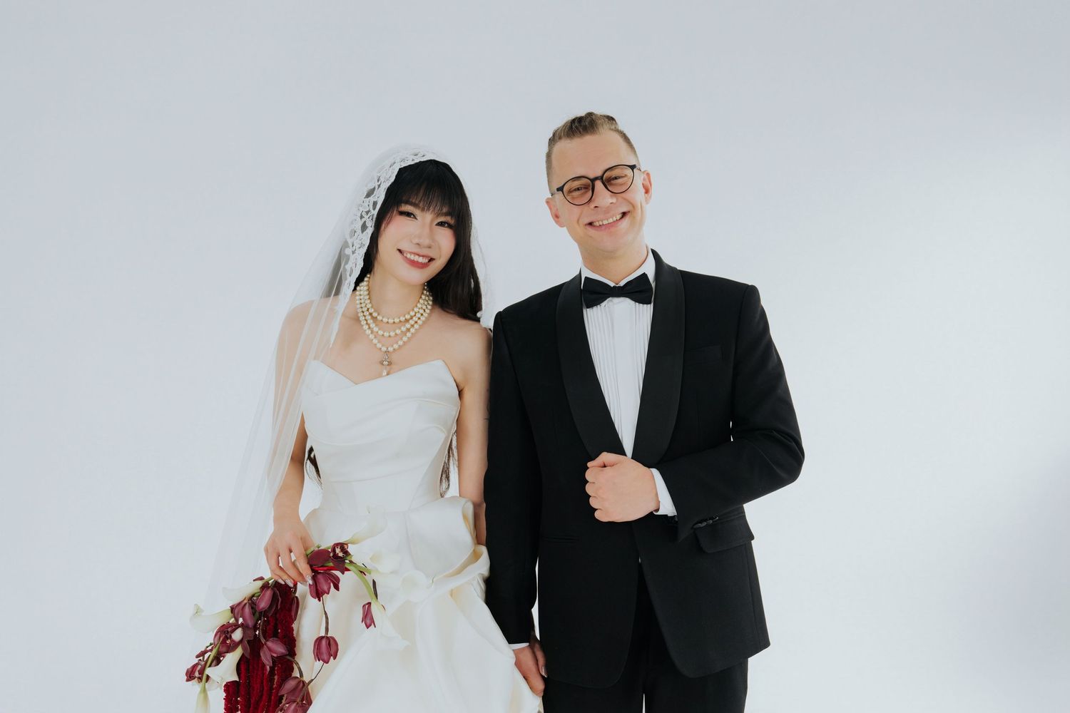 A bride in a white gown with veil holding red flowers stands beside a groom in a black tuxedo against a white background.
