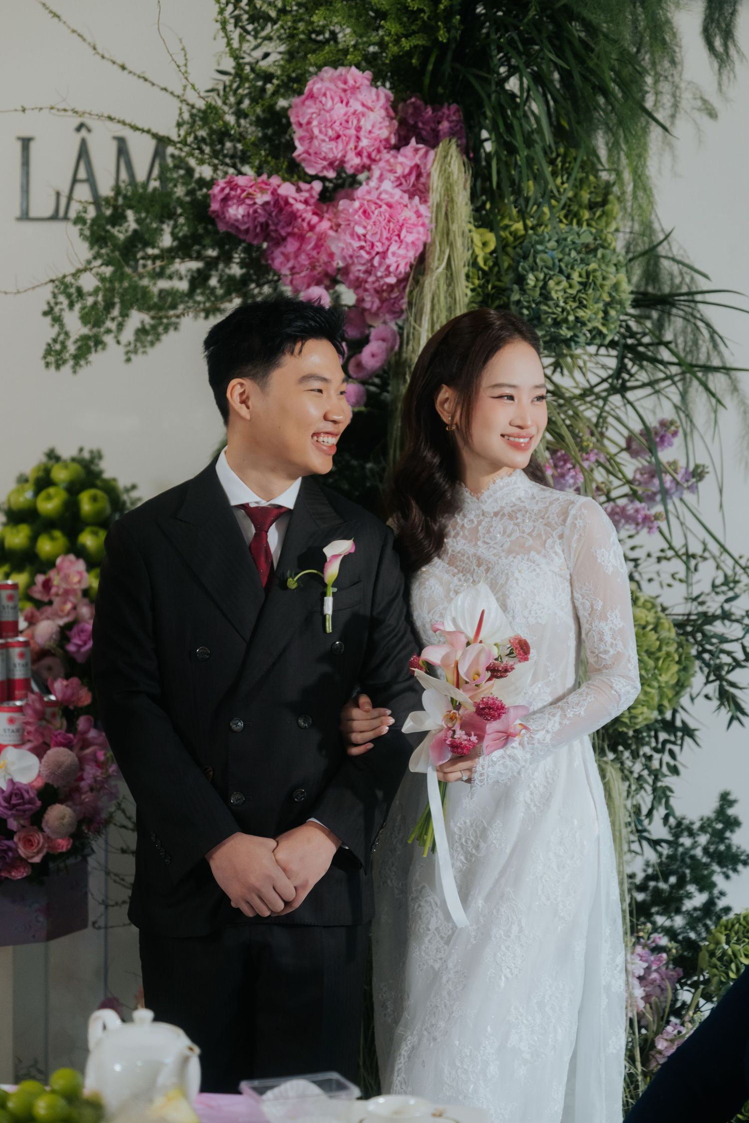 A couple in wedding attire smiles before a floral backdrop, the bride holding a bouquet in a white lace gown.