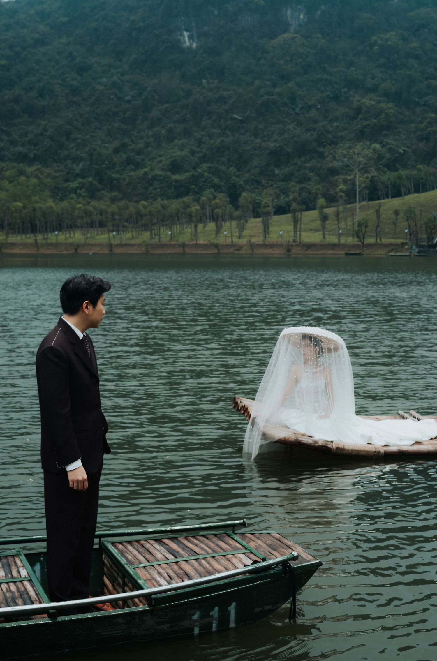 Groom in black suit stands on wooden boat gazing at veiled bride floating on water, with lush green hills behind.