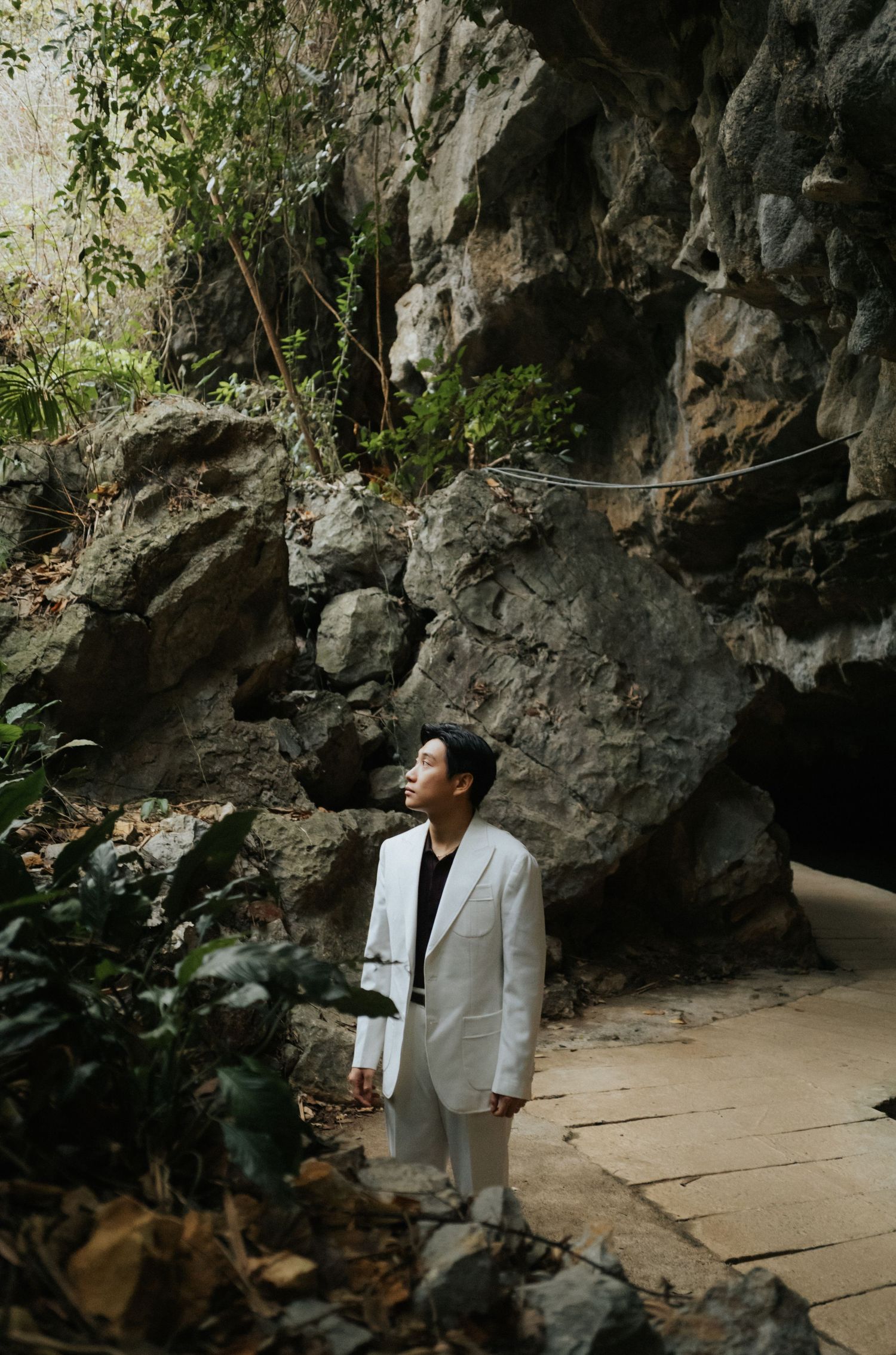 A man in a white suit stands gazing upward beside dramatic rocky cliffs and lush greenery on a stone path.