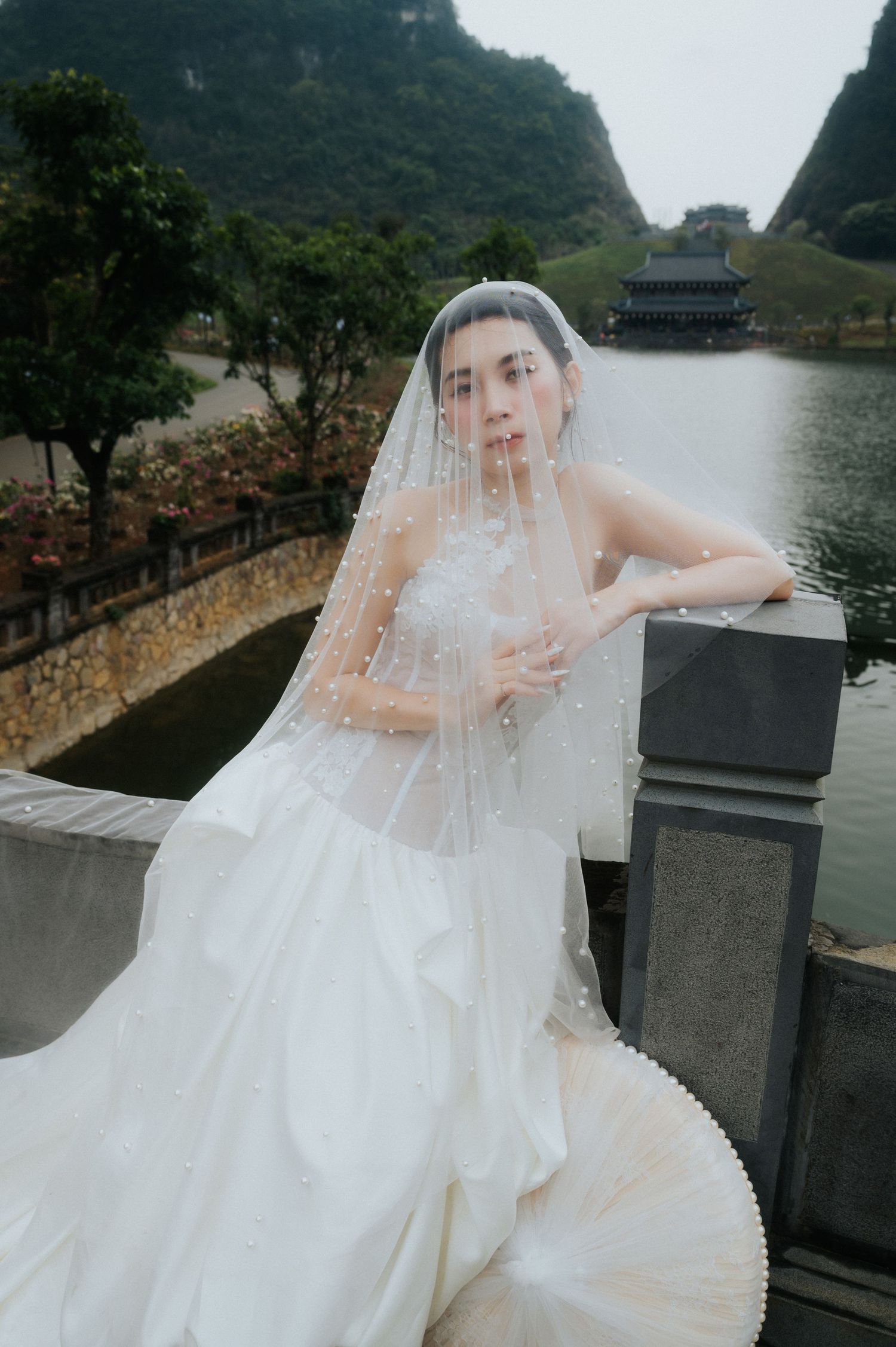 Bride in white wedding gown and pearl-adorned veil poses on stone bridge with misty karst mountains and lake backdrop.