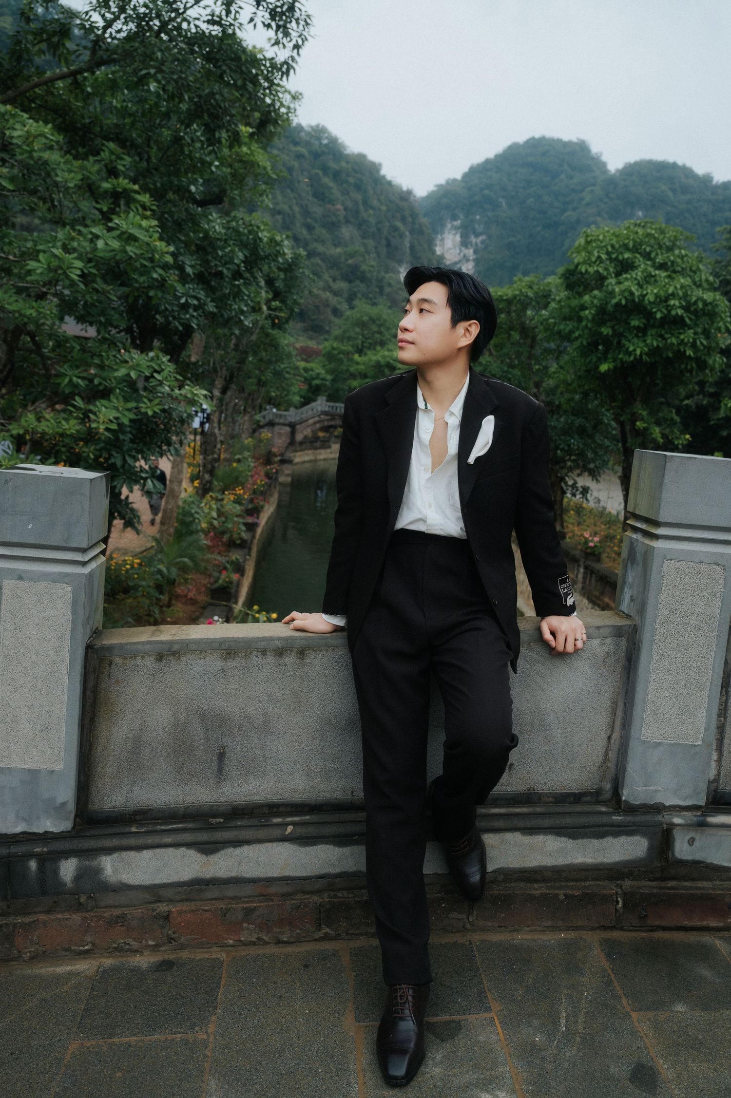 Young man in black suit leaning on stone railing with lush green mountains and trees in background.