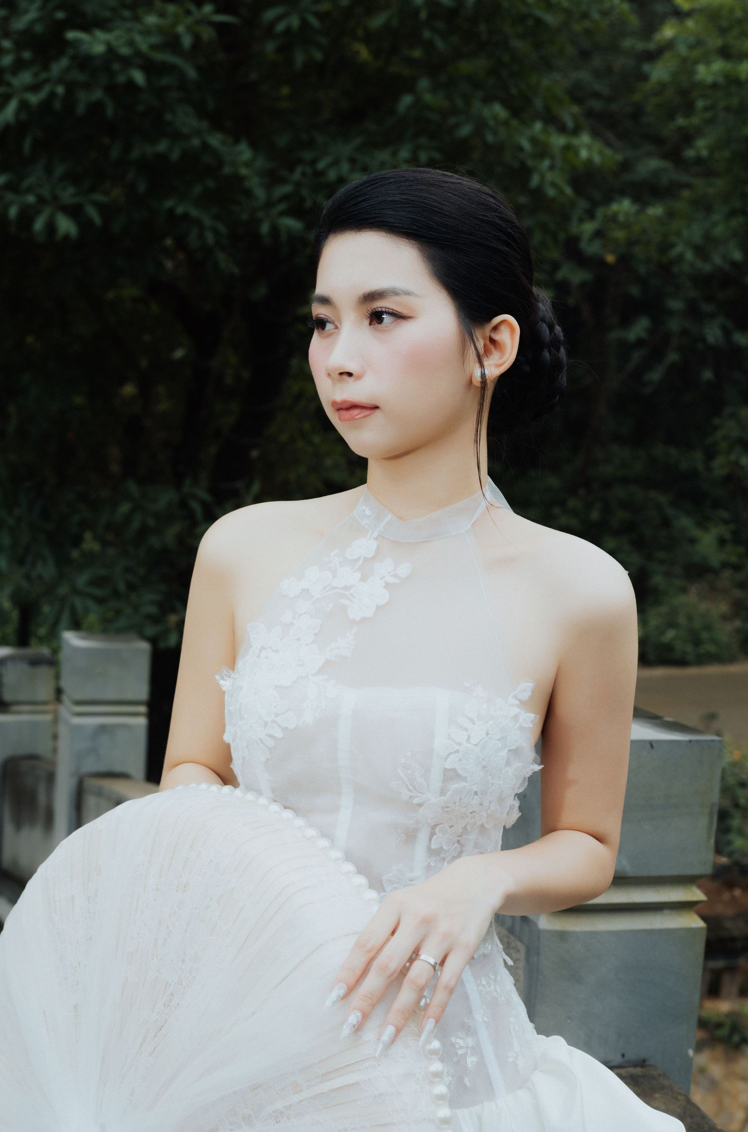 A young Asian woman in an elegant white lace halter gown poses gracefully outdoors with lush green foliage behind her.