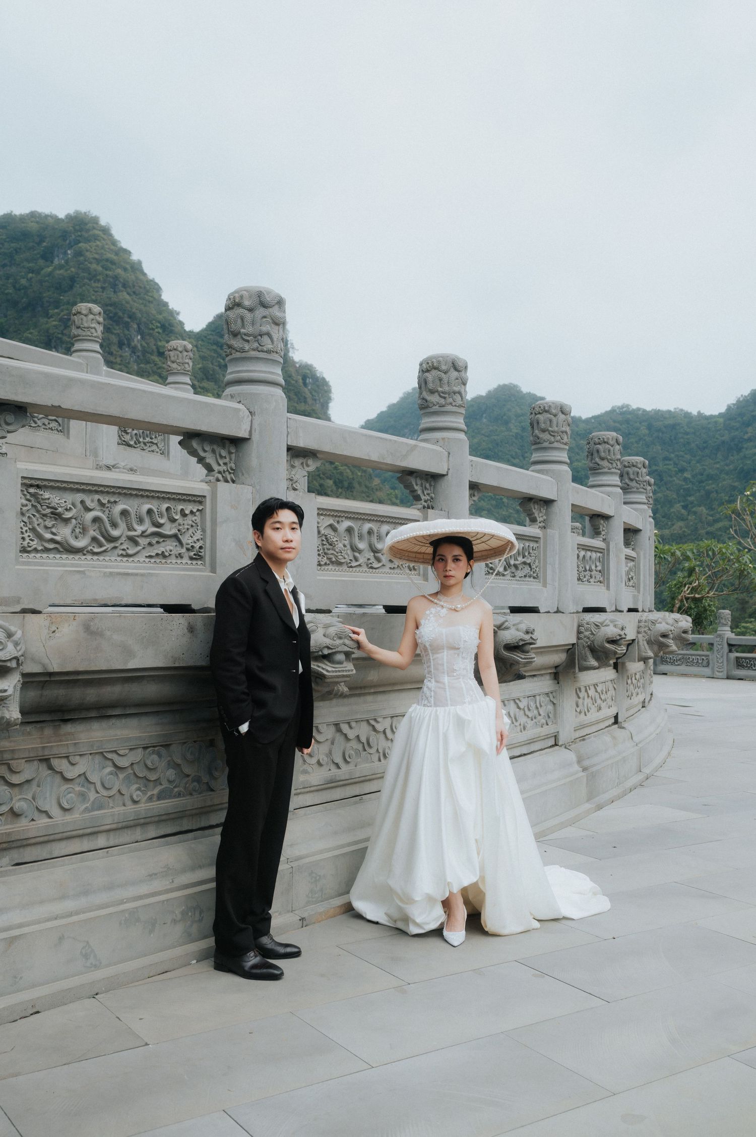 Couple in wedding attire posing by ornate stone railing with misty mountains backdrop at Asian temple.