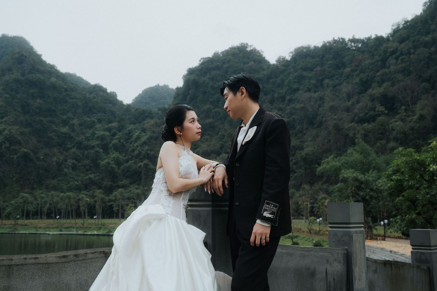 A couple in wedding attire poses romantically by a lakeside railing with lush green mountains in the background.