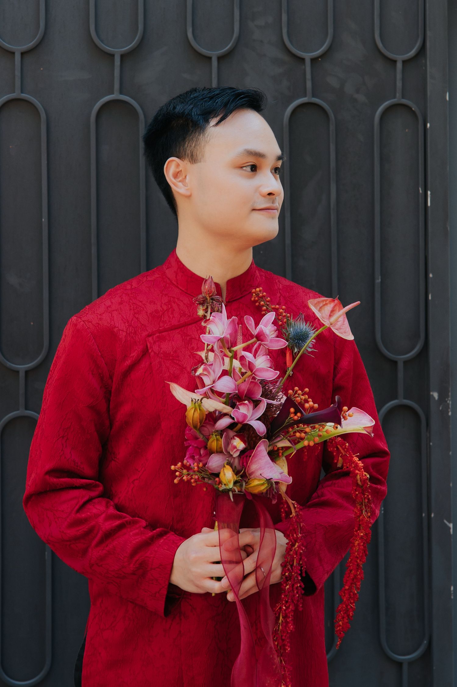 Young man in red Vietnamese ao dai holding a bouquet of pink and red flowers, standing against a dark metal gate.
