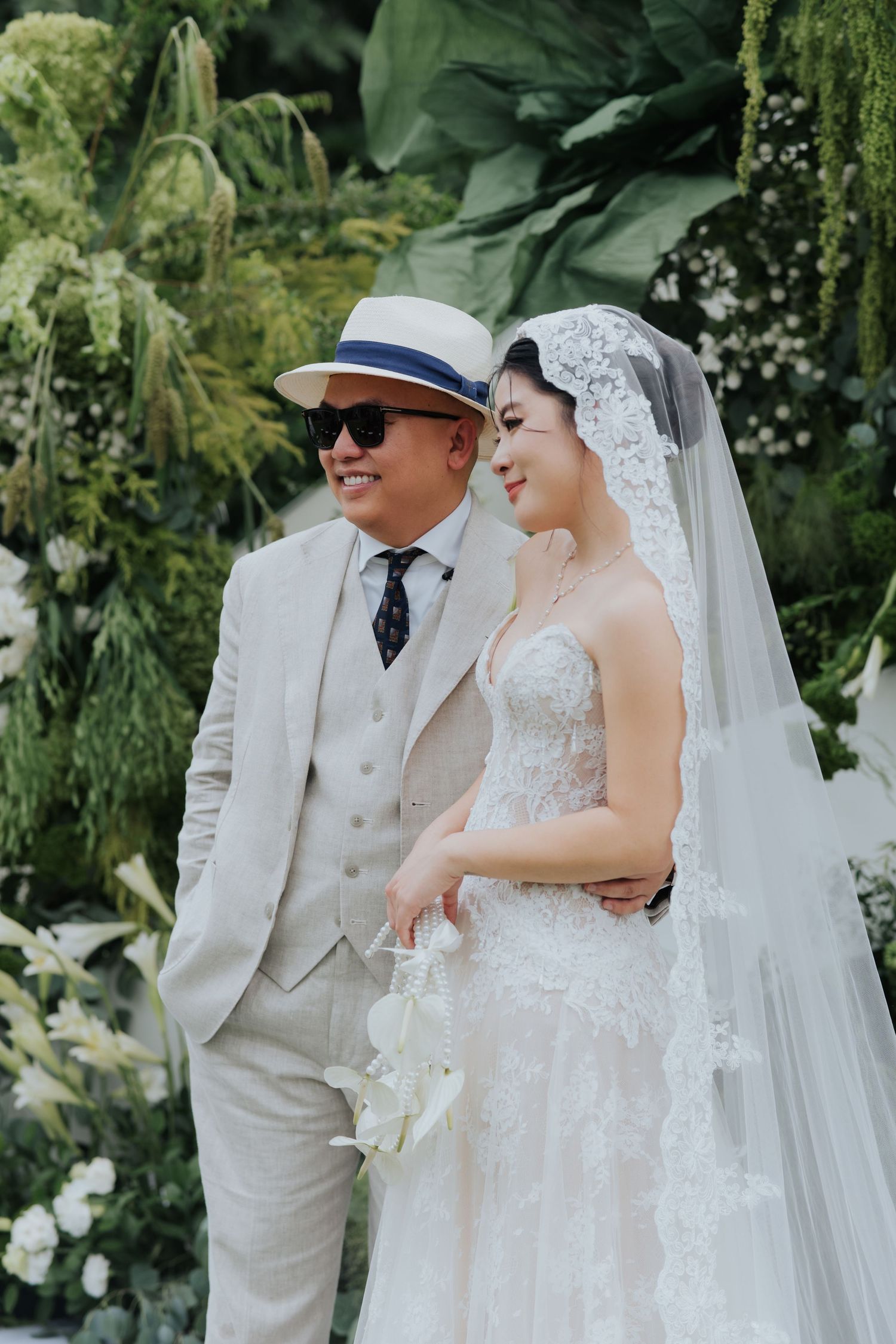 A bride in a lace wedding gown and veil stands with a groom in a cream suit and hat, surrounded by lush greenery.