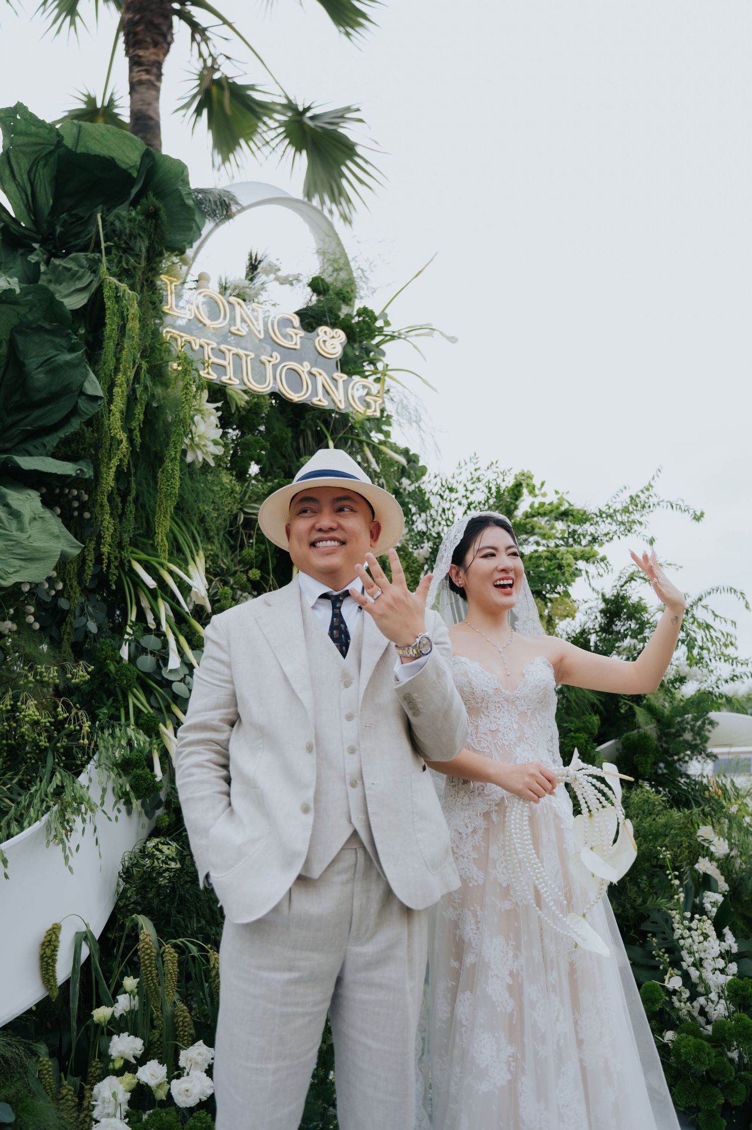 A joyful couple poses at their wedding ceremony, groom in white suit and hat, bride in lace gown, surrounded by lush greenery.