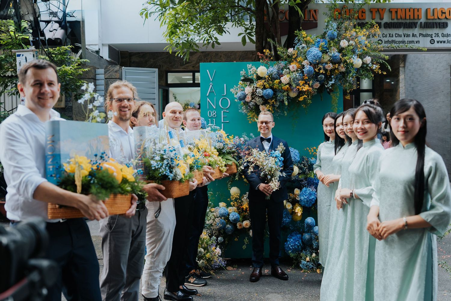 Group of people in formal attire celebrating outdoors with colorful floral arrangements at a Vietnamese event.