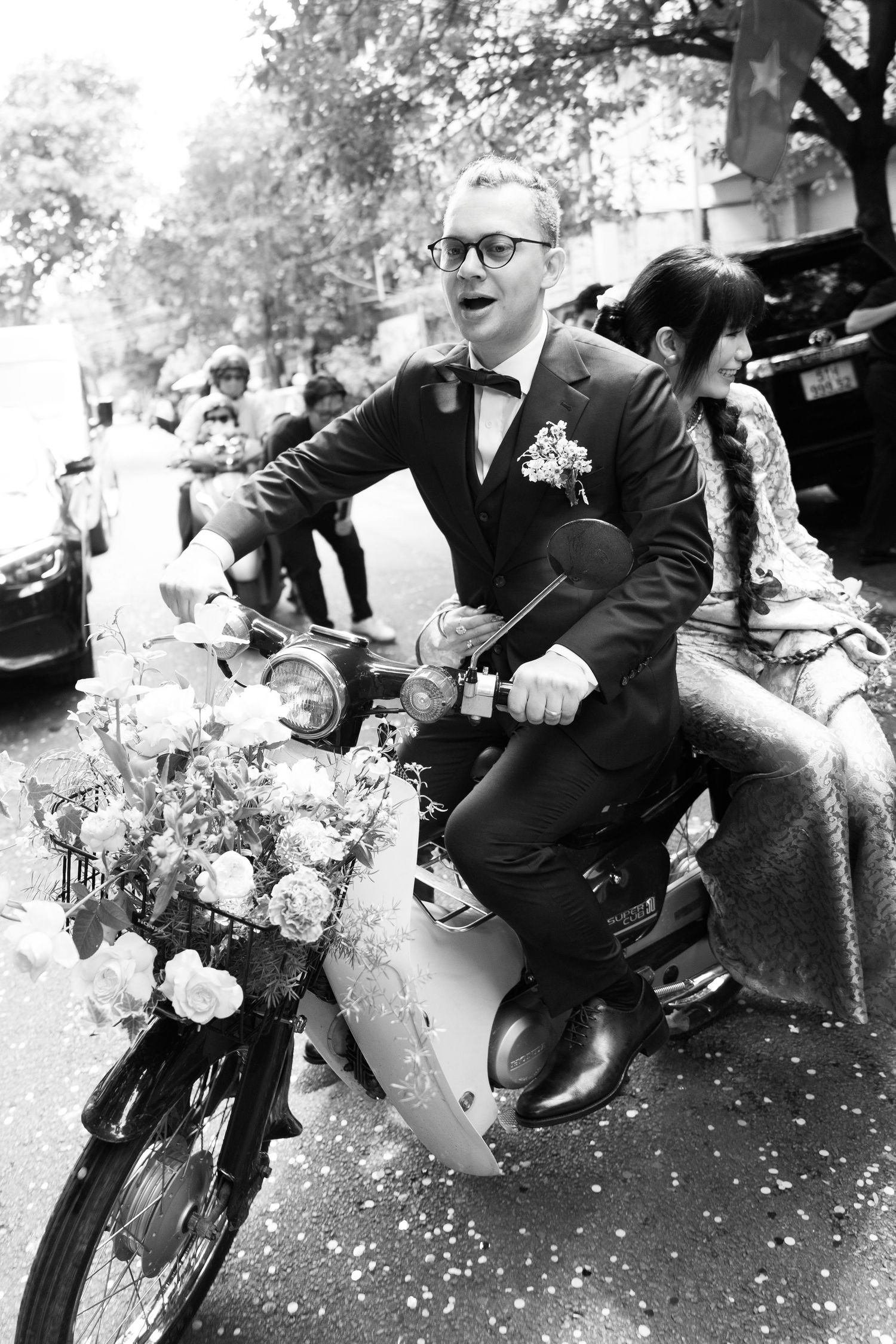 Groom in tuxedo rides flower-adorned motorcycle with bride during wedding celebration, black and white photo.