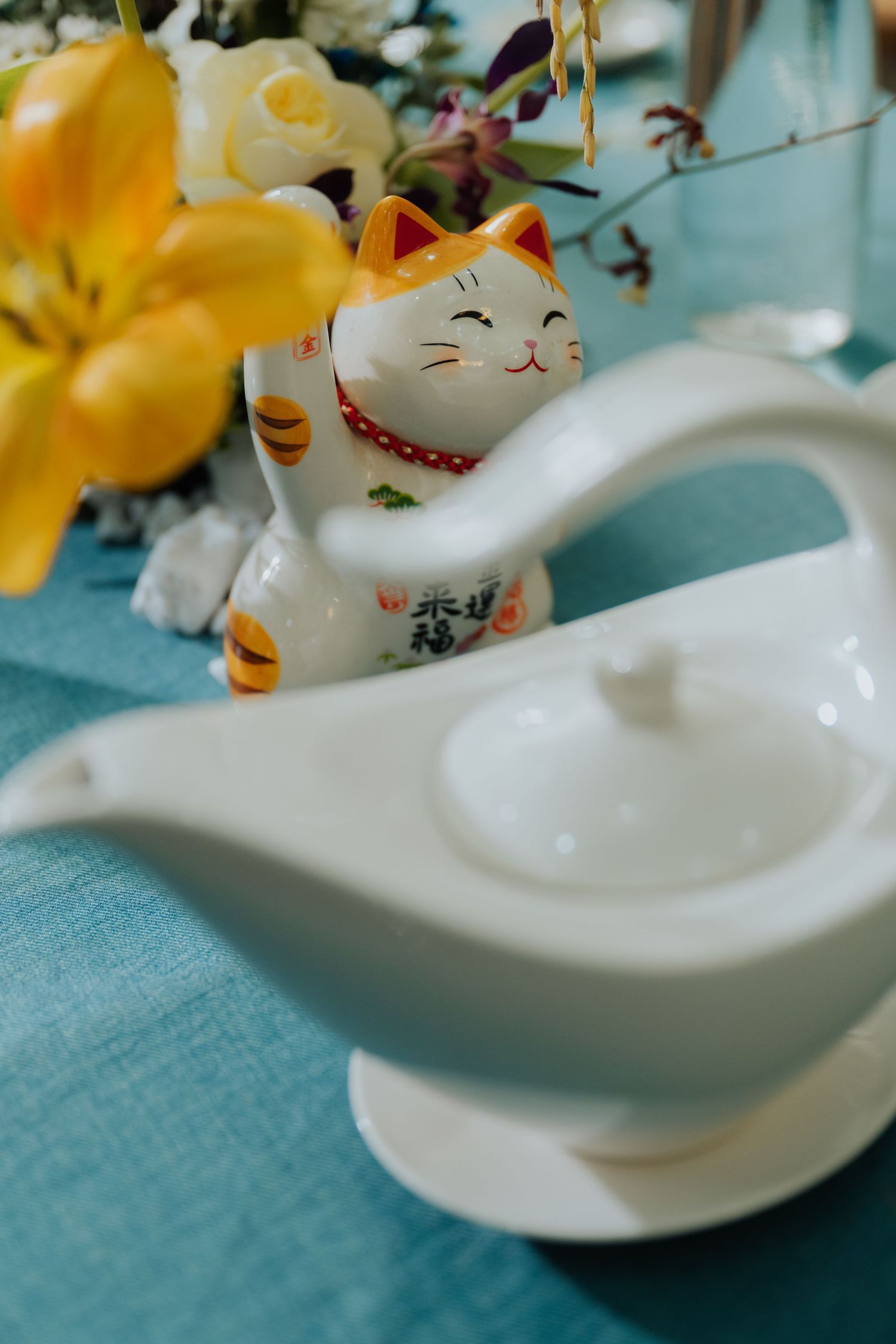 White ceramic gravy boat and lucky cat figurine on teal tablecloth with yellow flowers in background.