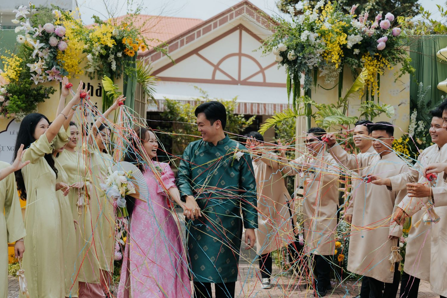Vietnamese wedding couple in traditional ao dai attire walk through a floral arch as guests celebrate outdoors.