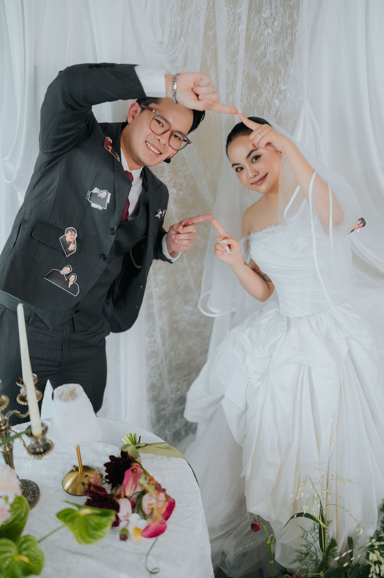 A smiling couple in wedding attire poses making a heart shape with their hands at a decorated reception table.