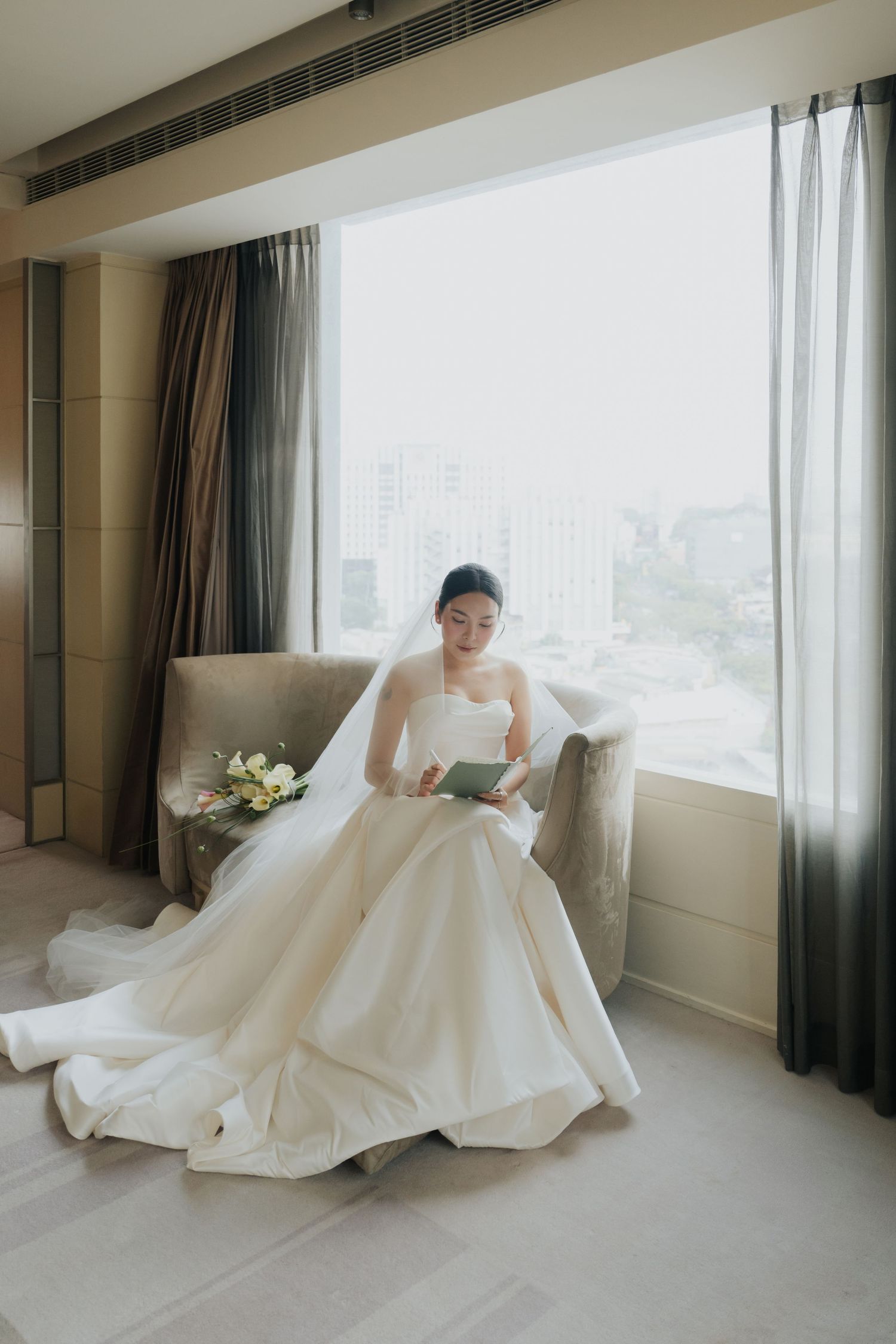 Bride in a white ball gown and veil reads a card while seated by a large window with city views.