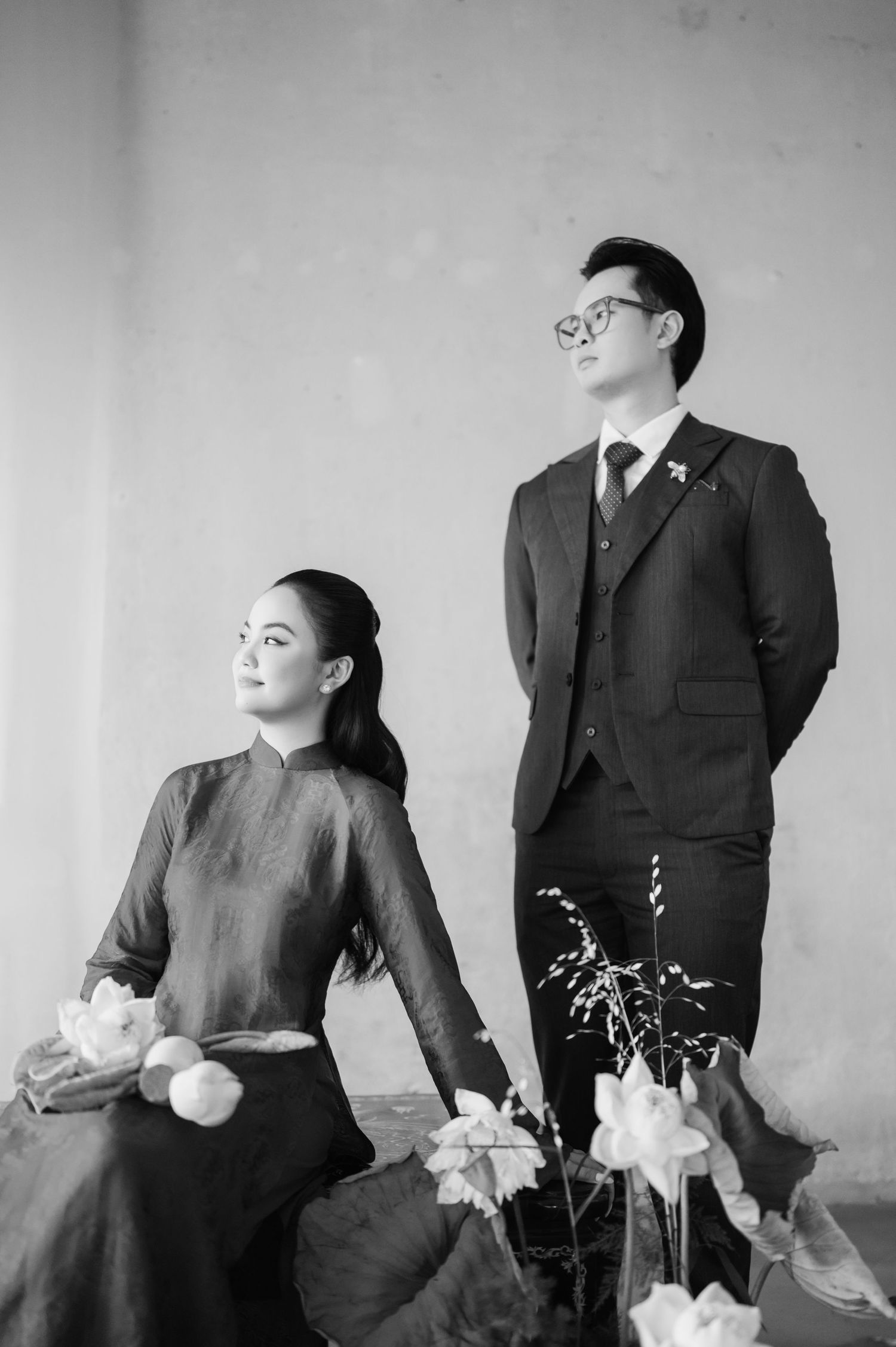 Black and white portrait of an elegant couple, woman in traditional ao dai holding lotus flowers, man in suit standing beside her.
