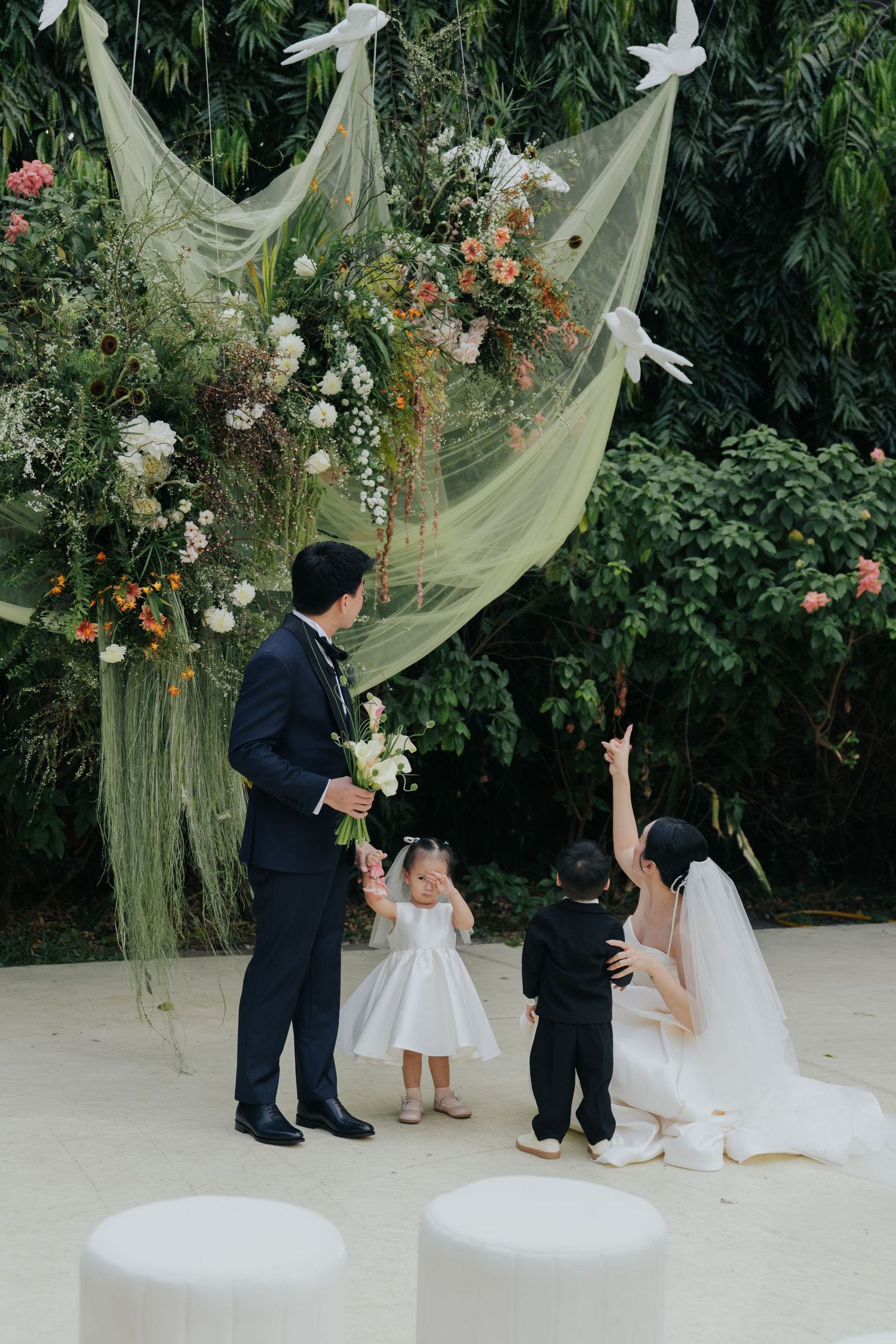 Bride and groom with flower girl and ring bearer at outdoor wedding ceremony with floral arch and green draping.