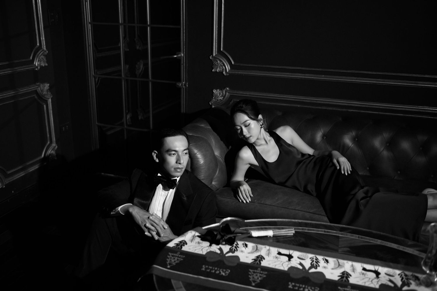 Elegant couple in black tie attire lounging on a leather sofa in a moody black and white portrait.