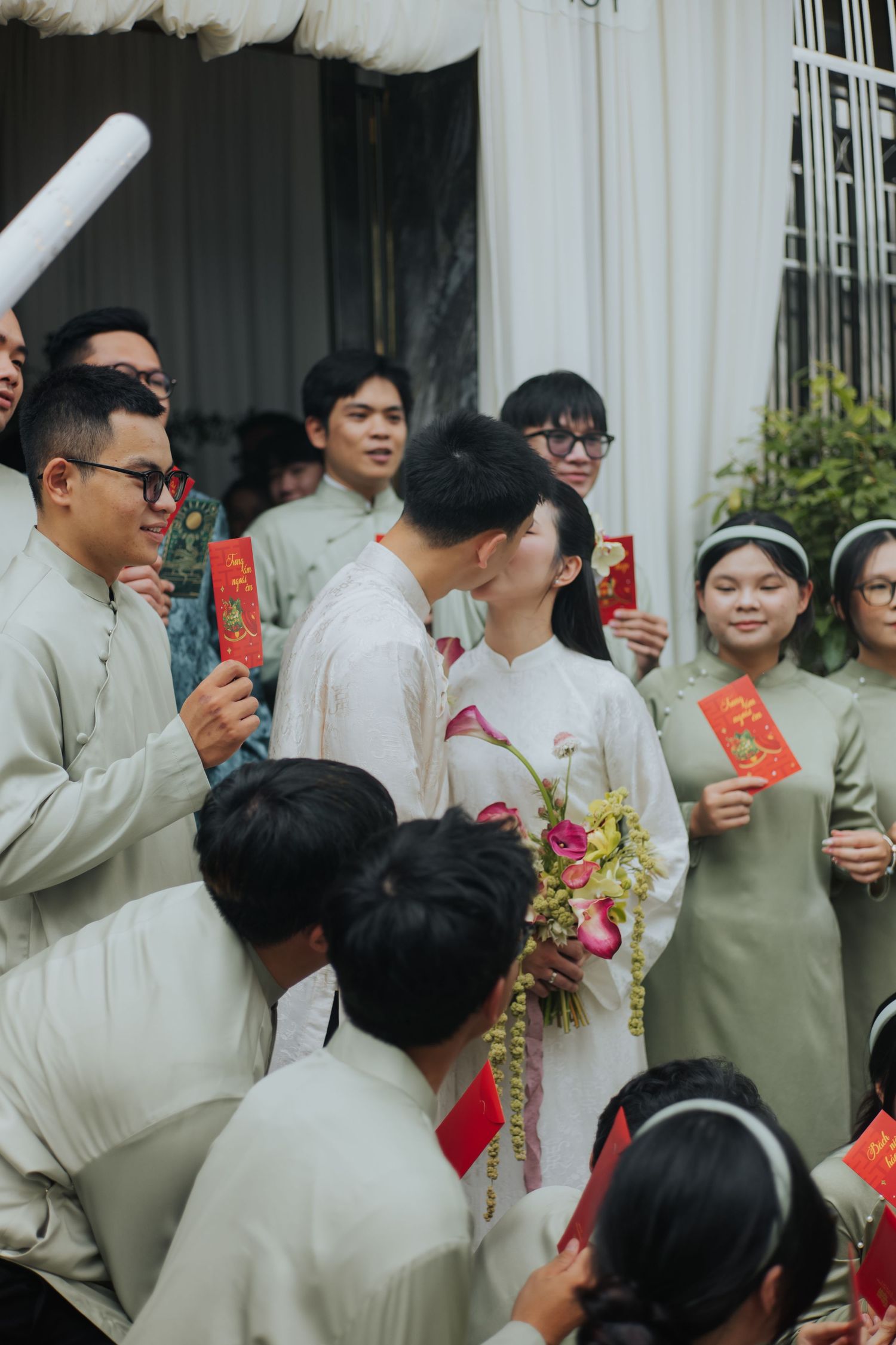 Vietnamese wedding ceremony with couple kissing, guests in traditional ao dai holding red envelopes outdoors.