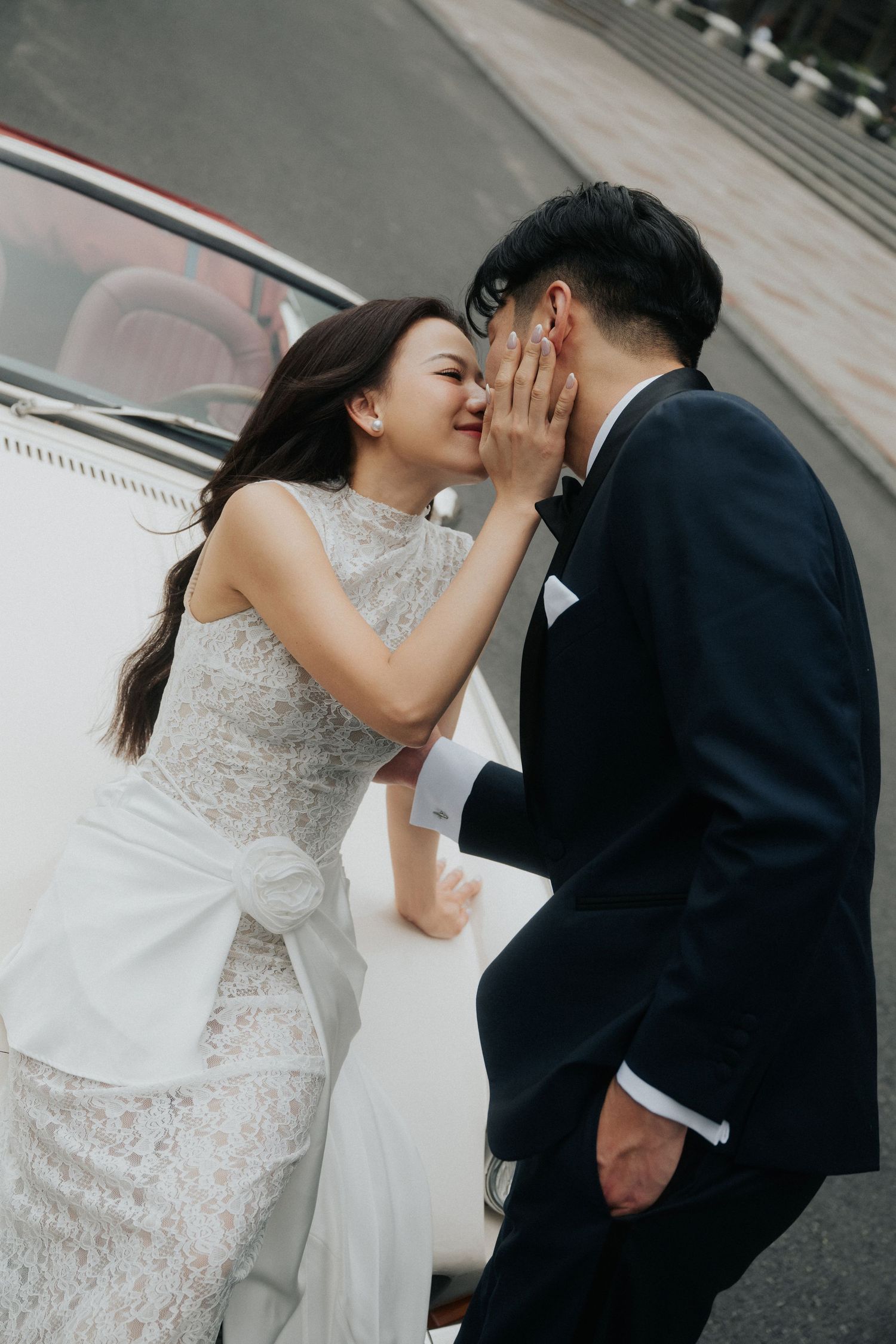 A bride in a lace gown tenderly touches her groom's face as they share an intimate moment beside a white vintage car.