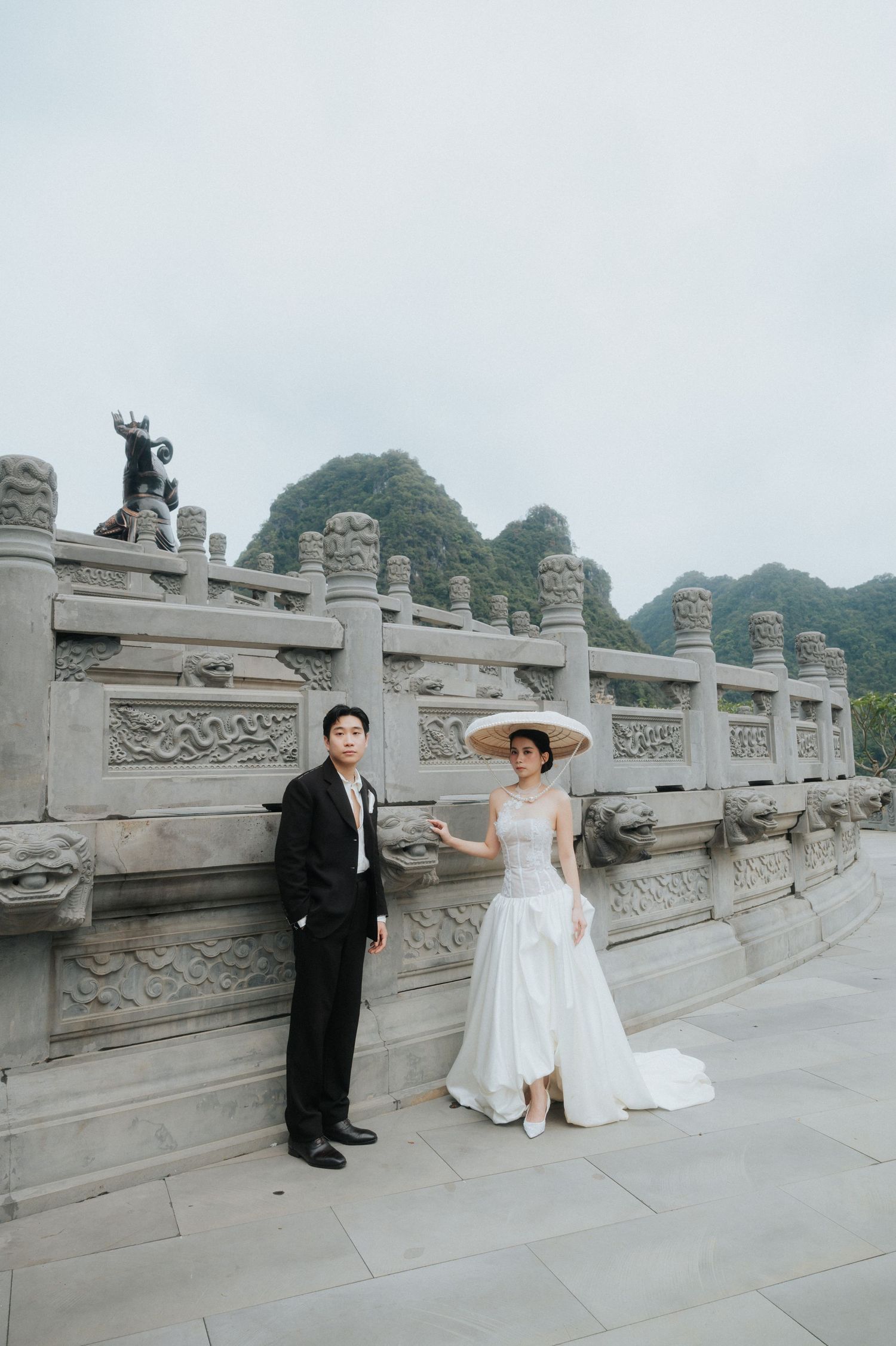 Wedding couple posing at carved stone railing with karst mountains backdrop, bride in white gown and straw hat.