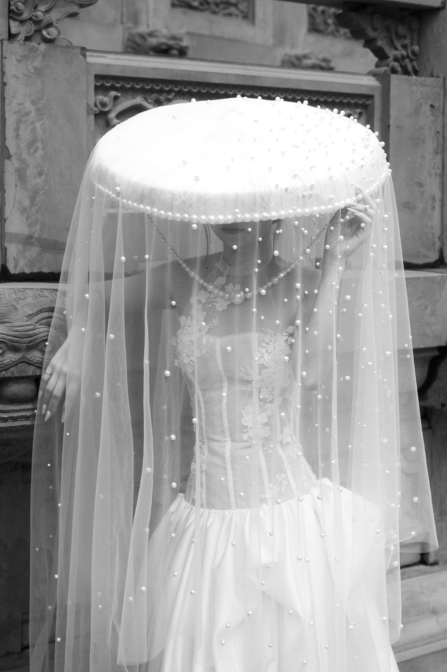 Bride in white lace wedding gown wearing pearl-adorned veil covering face, black and white photo.