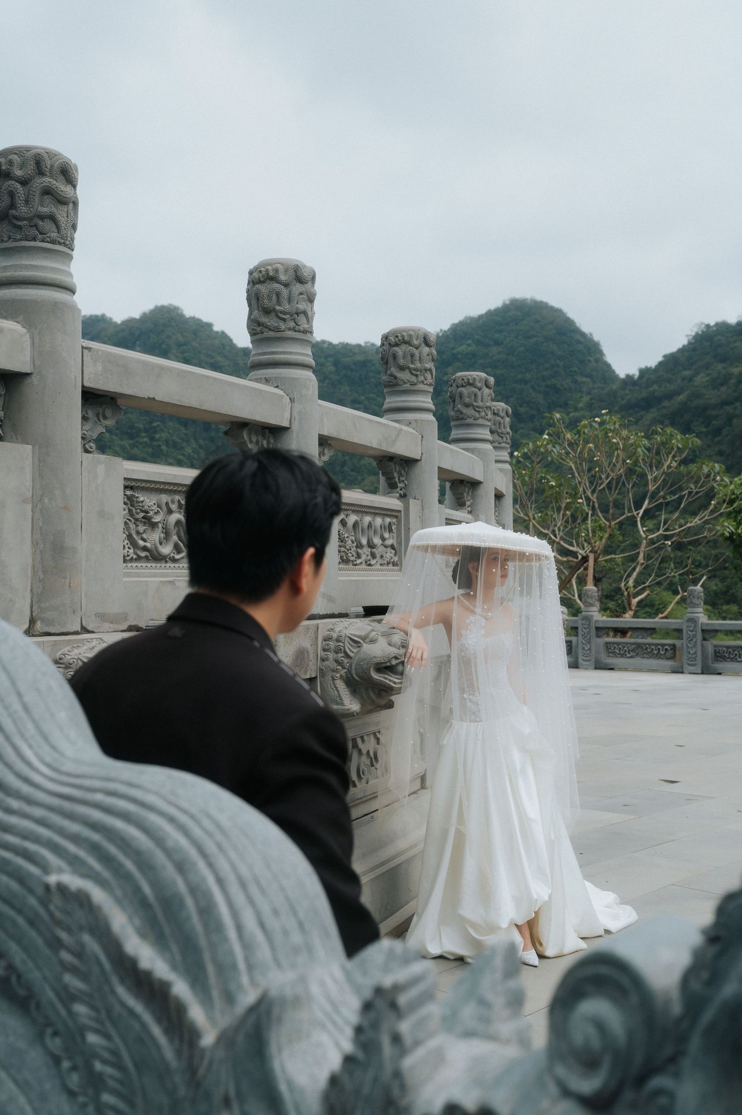 A bride in white gown and veil holds umbrella near ornate stone balustrade with misty mountains backdrop.