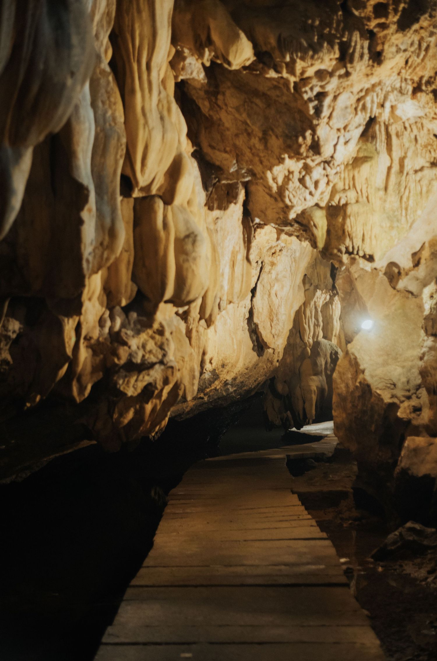 Wooden boardwalk path through illuminated limestone cave with stalactites and glowing rock formations.