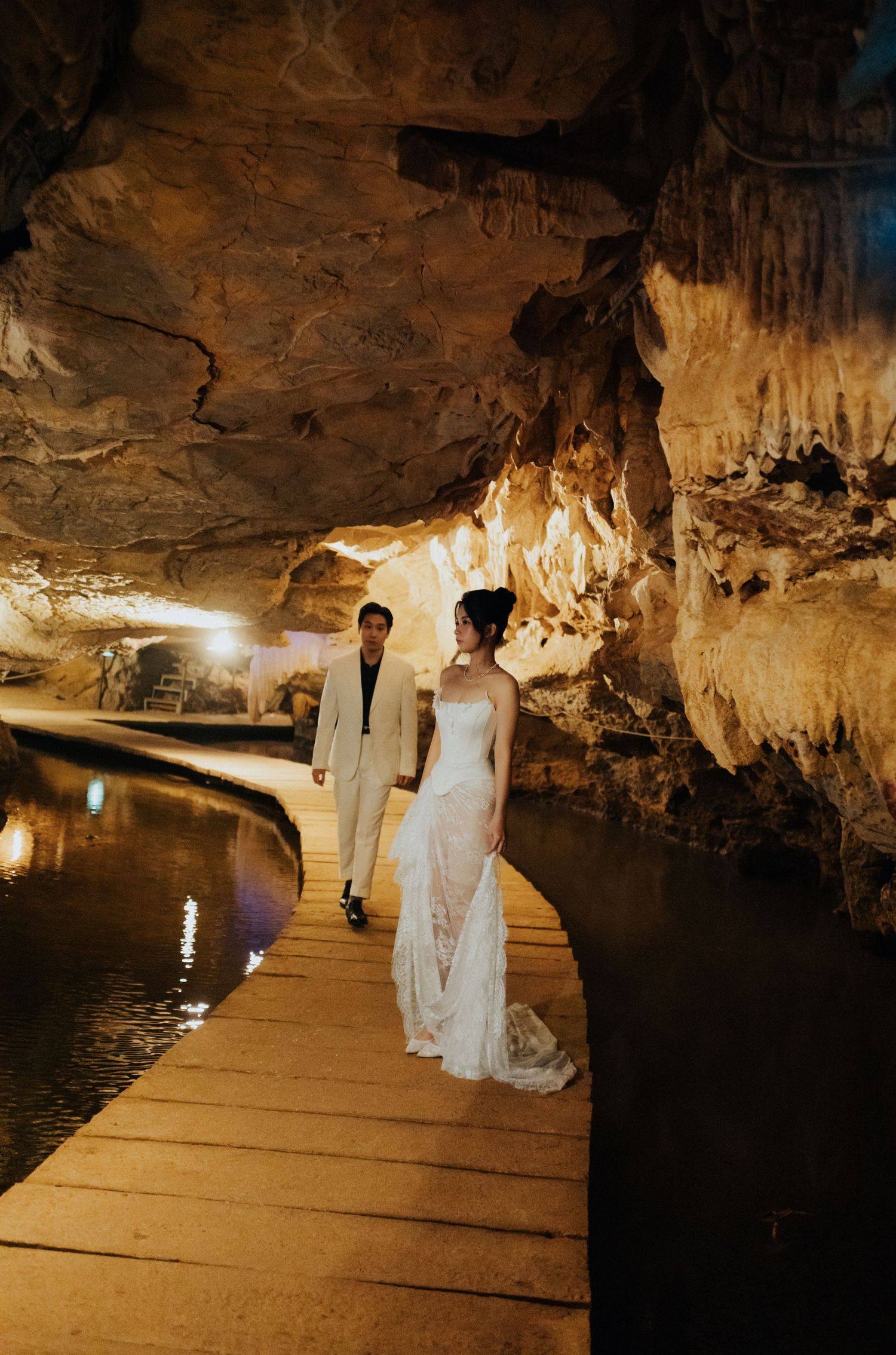 Bride and groom walk hand-in-hand on a wooden boardwalk inside a stunning cave with stalactites and glowing lights.