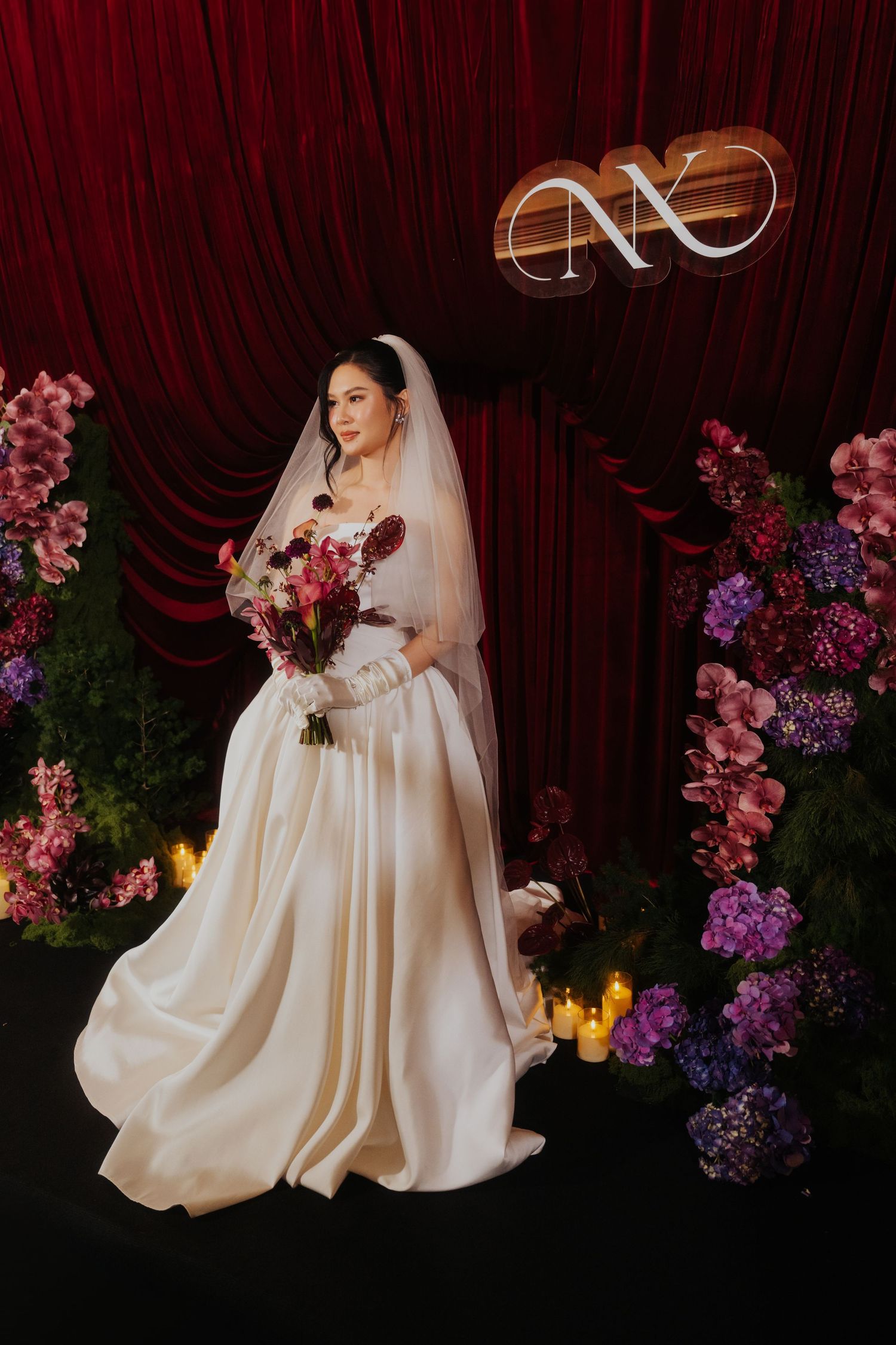 Bride in white satin gown and veil holds colorful bouquet before red drape backdrop with floral arrangements and candles.