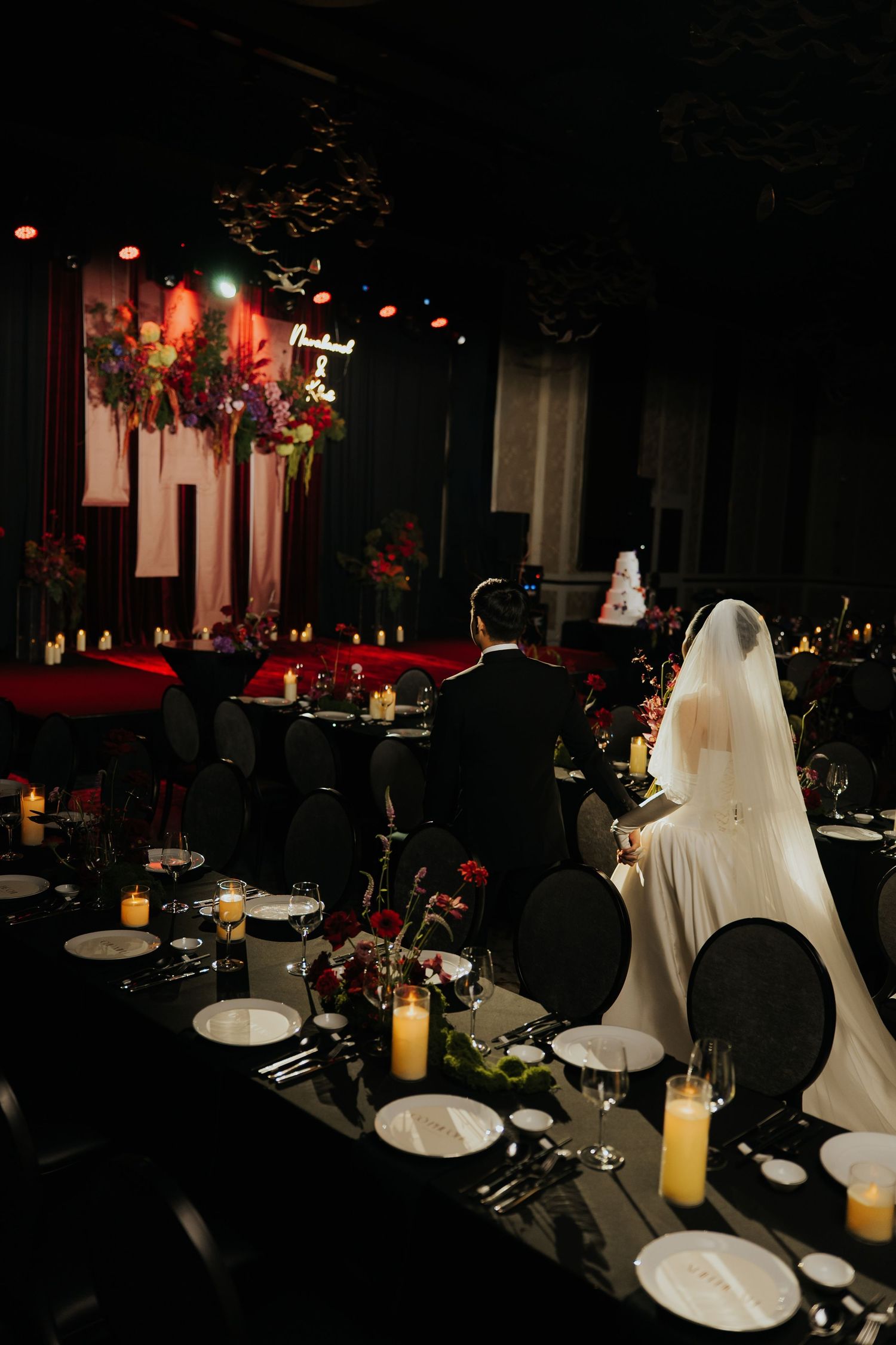 Elegant wedding reception with bride and groom at candlelit black tables, dramatic red floral backdrop, and moody dark decor.