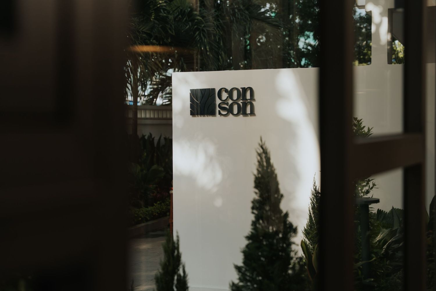 White signage displaying the Coin Son logo surrounded by lush green trees, viewed through a wooden fence.