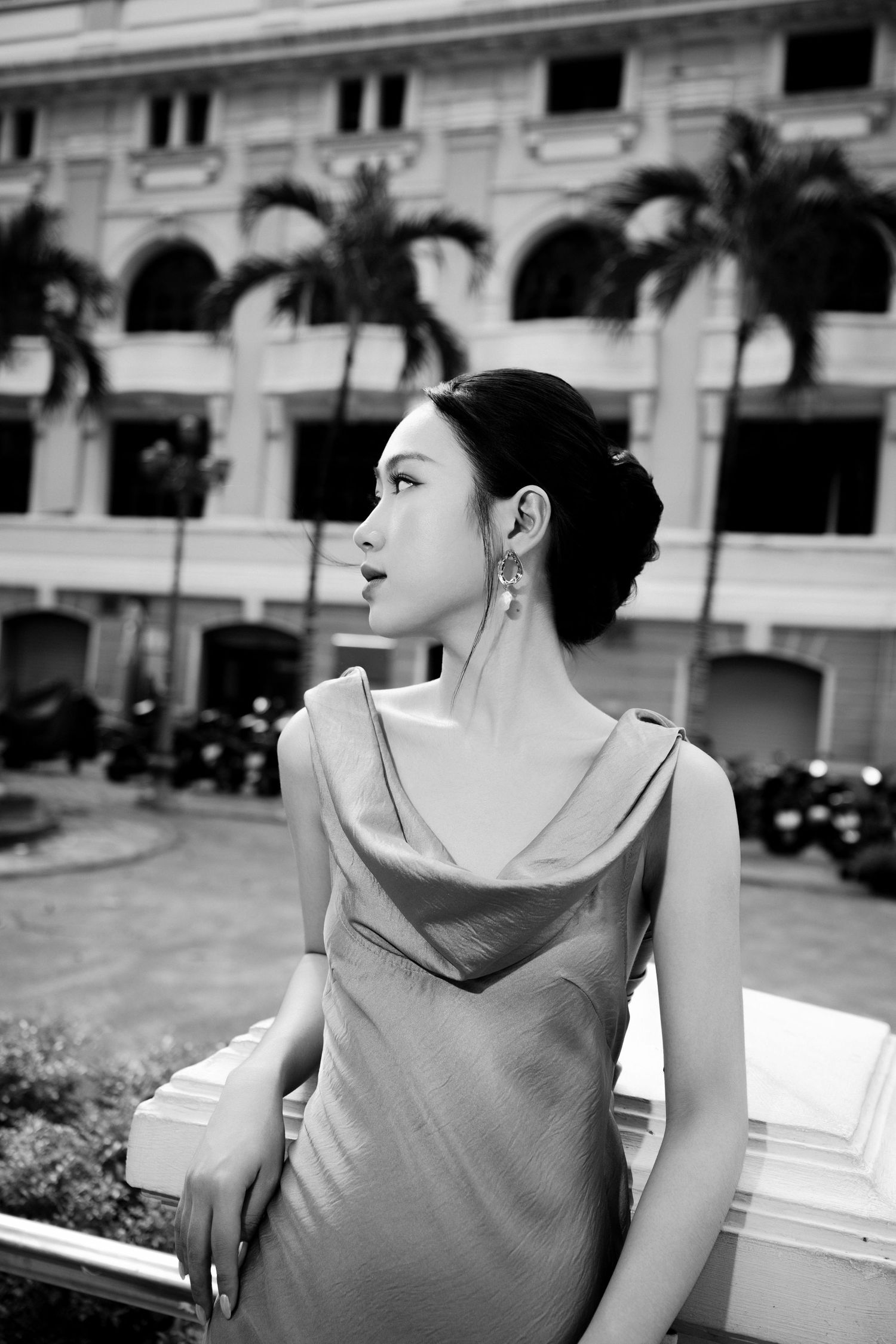Black and white photo of an elegant Asian woman in a satin dress, posing outdoors near a classic building with palm trees.