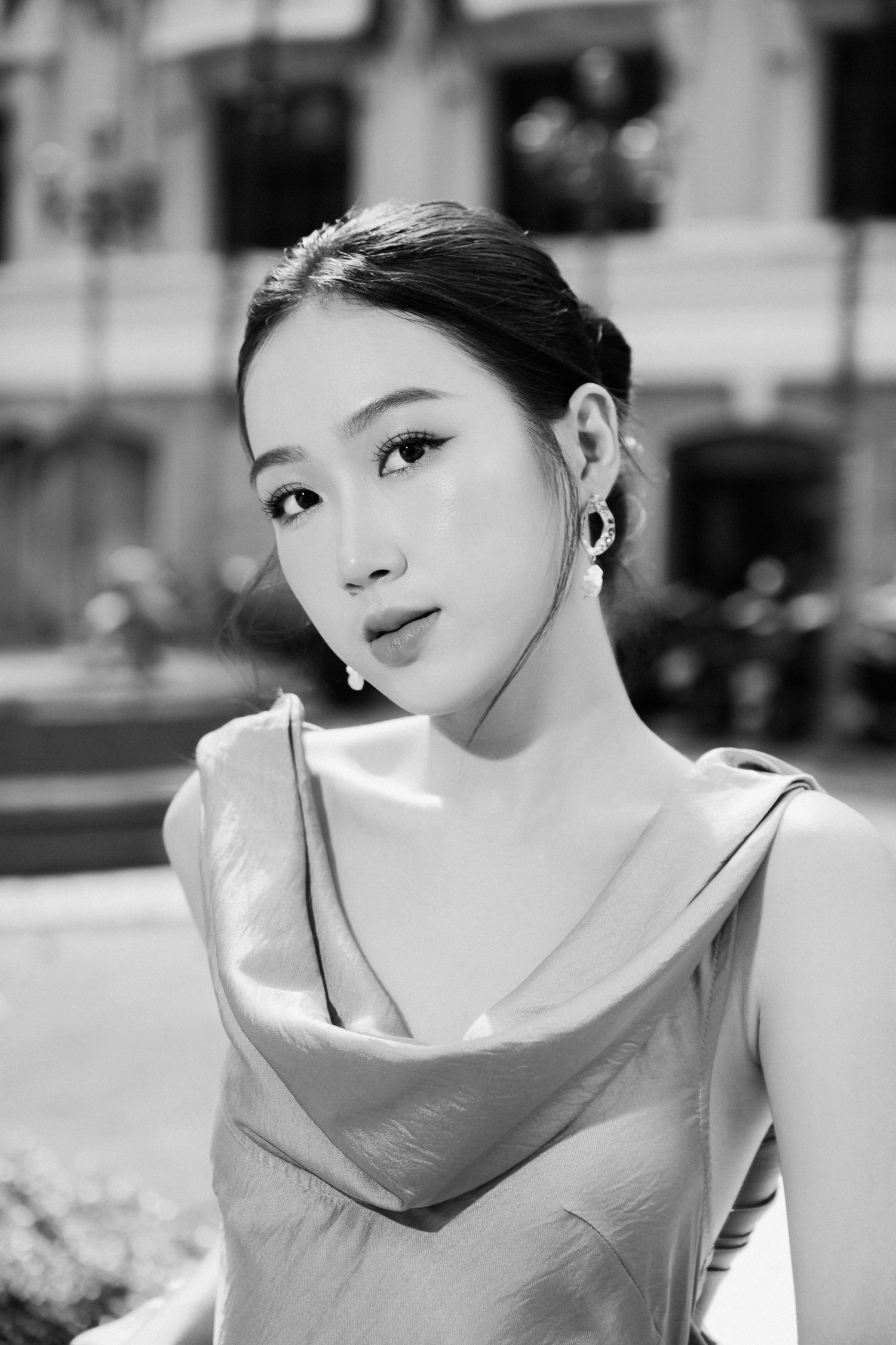 Black and white portrait of an elegant young Asian woman in a satin dress with hoop earrings outdoors.
