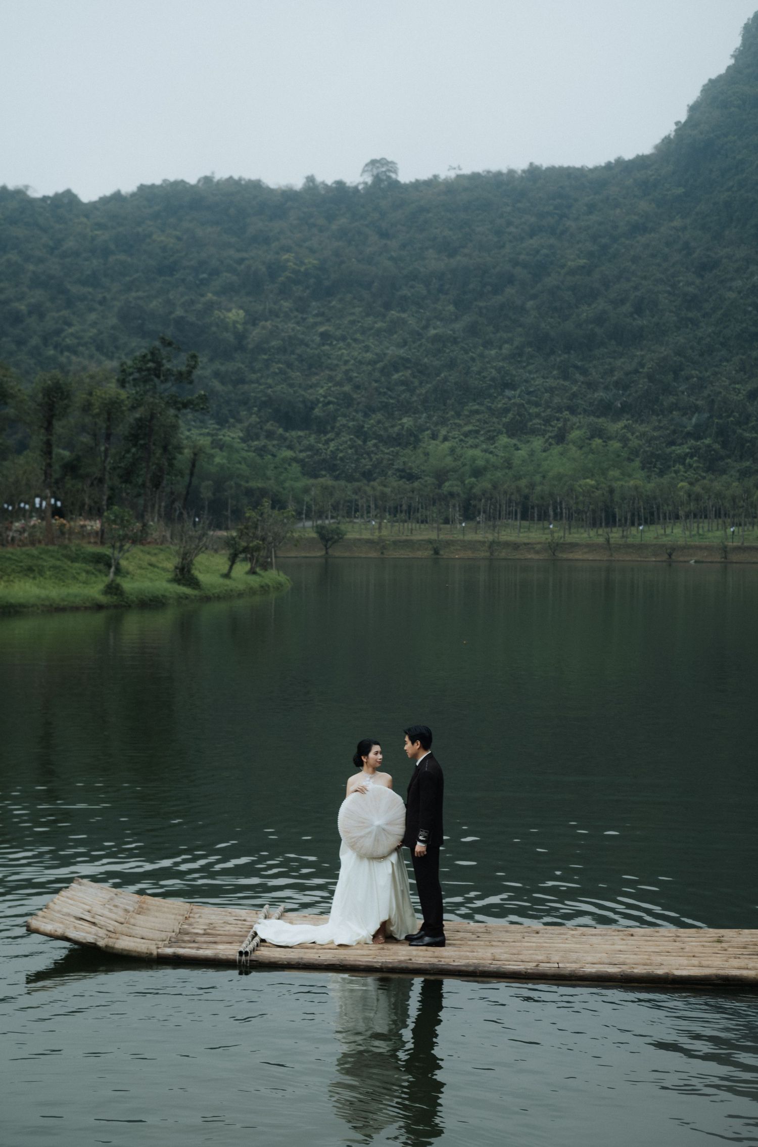 A bride and groom stand on a bamboo raft over a calm lake surrounded by lush green mountains.