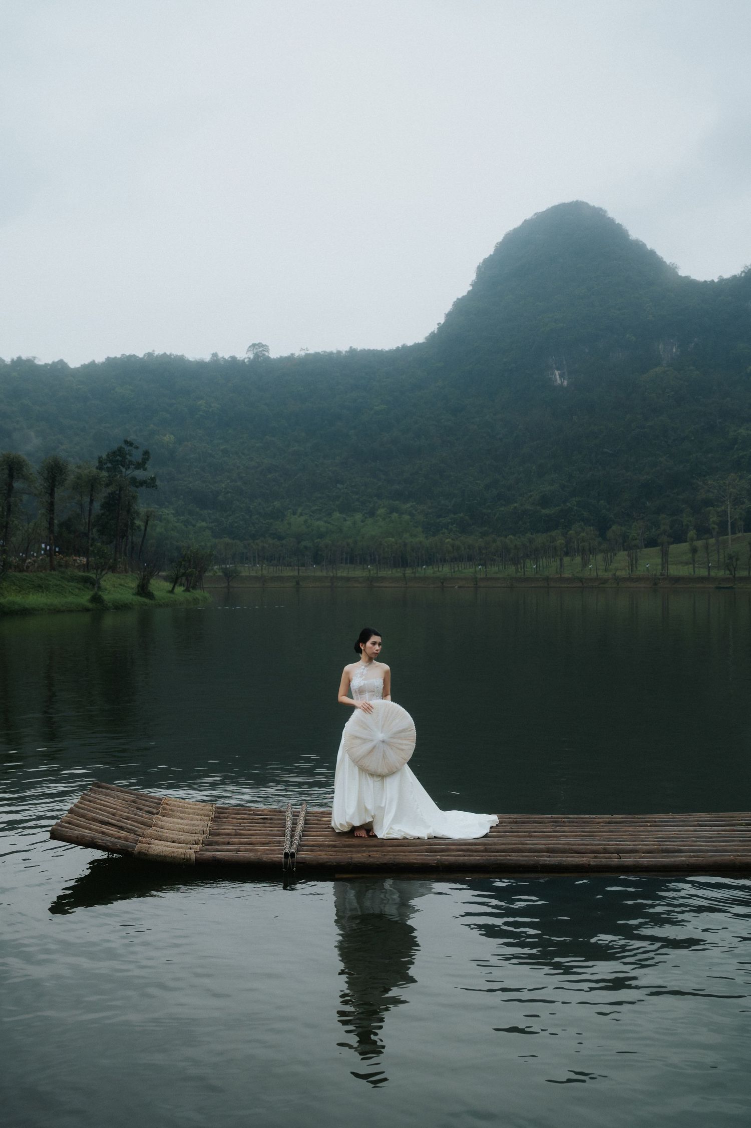 Bride in white gown holding a large hat stands on a bamboo dock over a serene lake with misty green mountains.