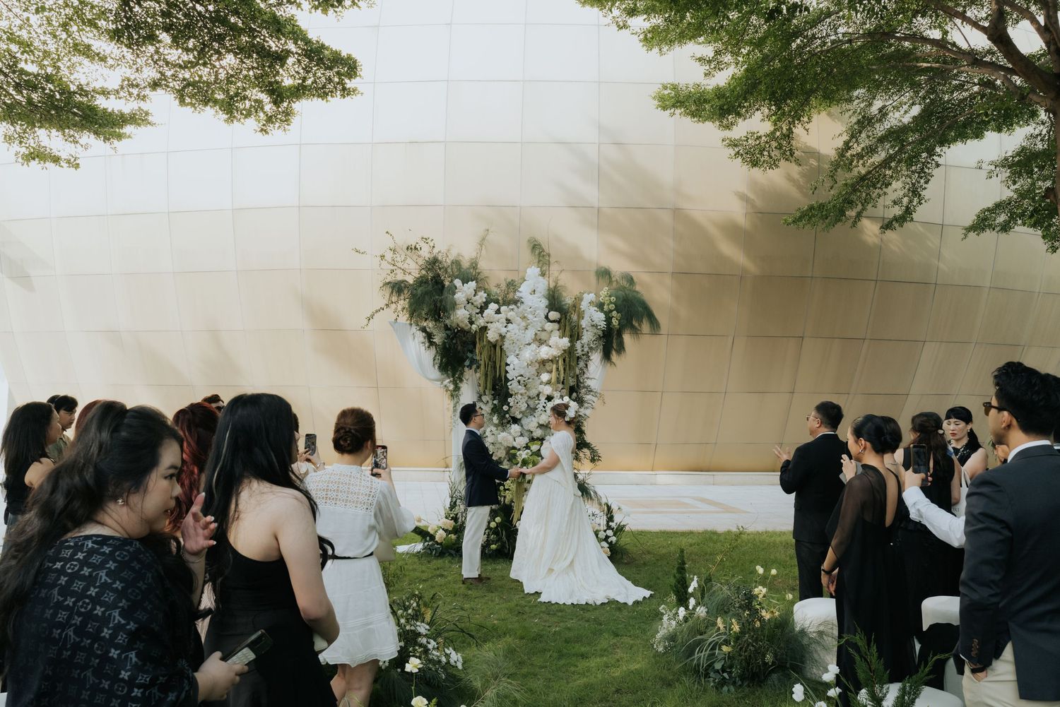 Outdoor wedding ceremony on green lawn with floral arch, bride and groom, and guests gathered beside a modern golden building.