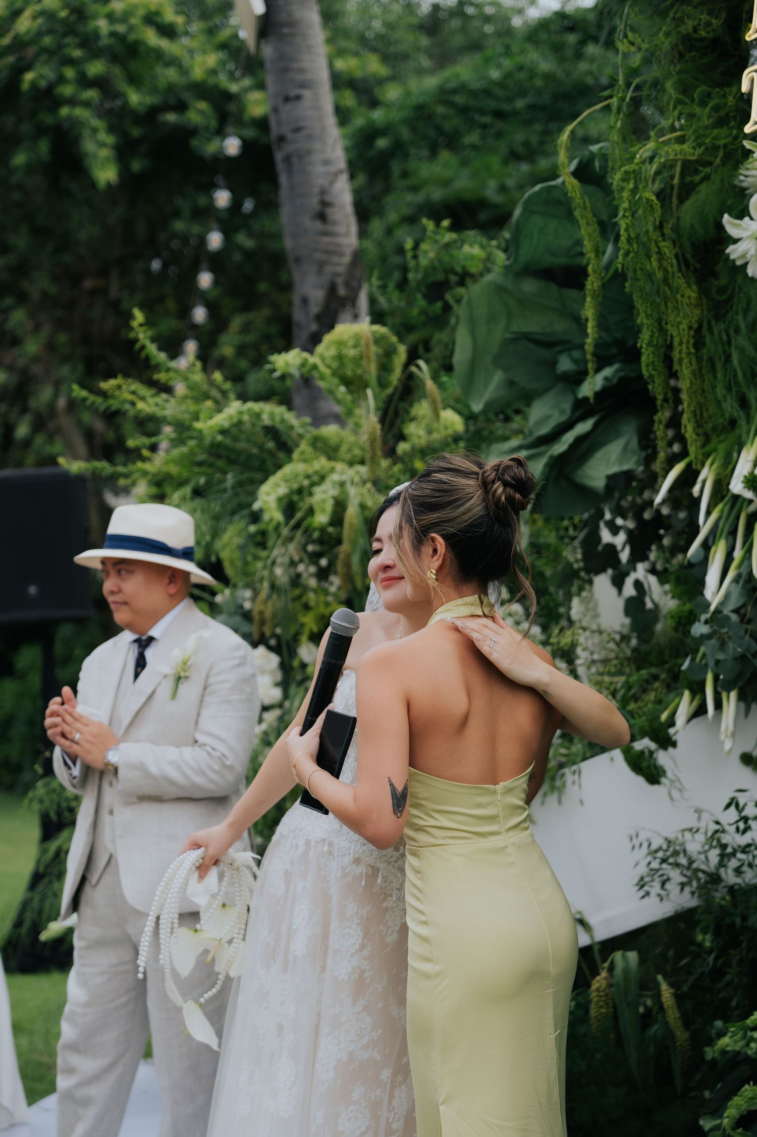 Bride in white gown and bridesmaid in green halter dress share a joyful moment at an outdoor tropical wedding ceremony.