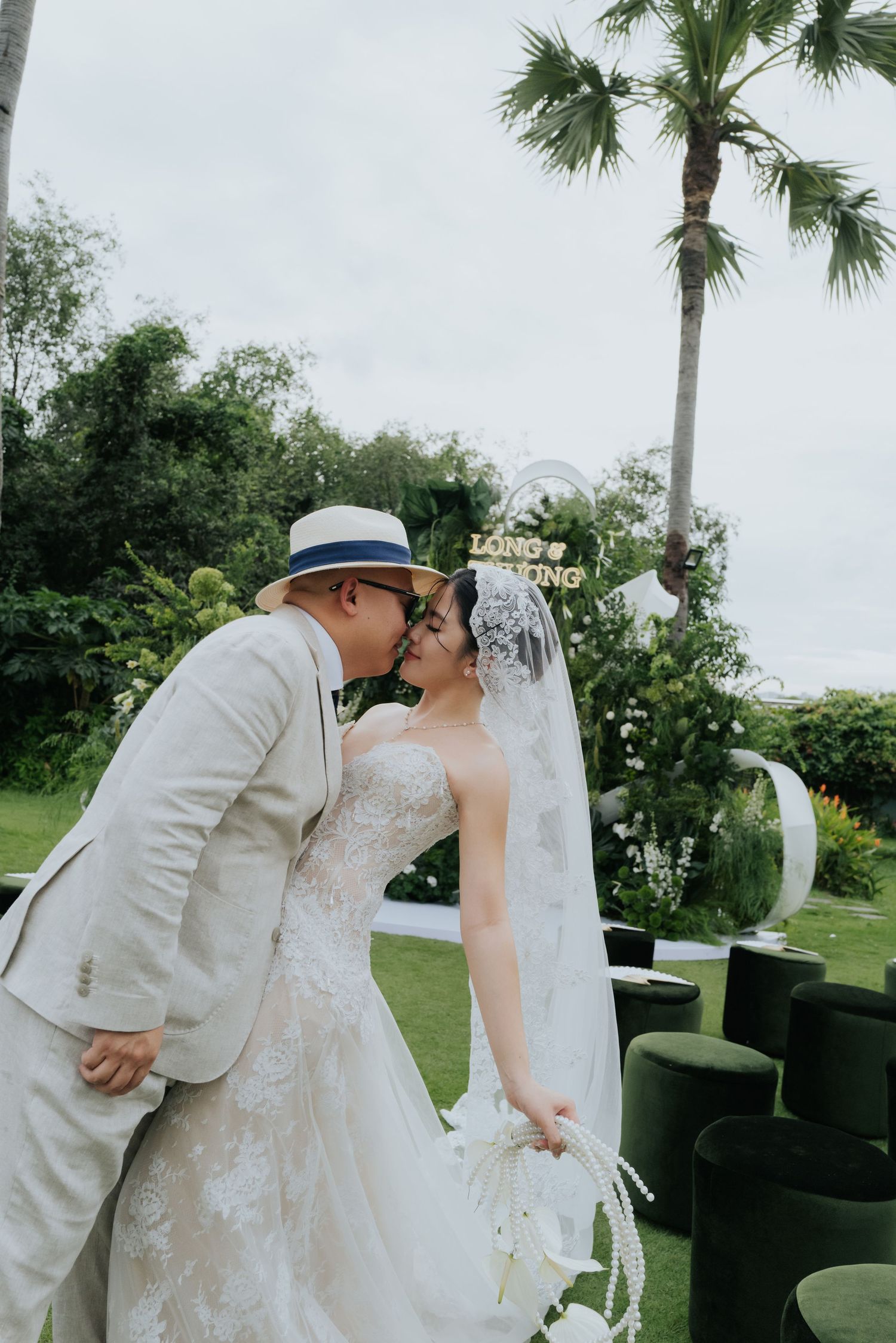 A couple shares a kiss at an outdoor wedding ceremony, groom in beige suit and hat, bride in lace gown with veil.
