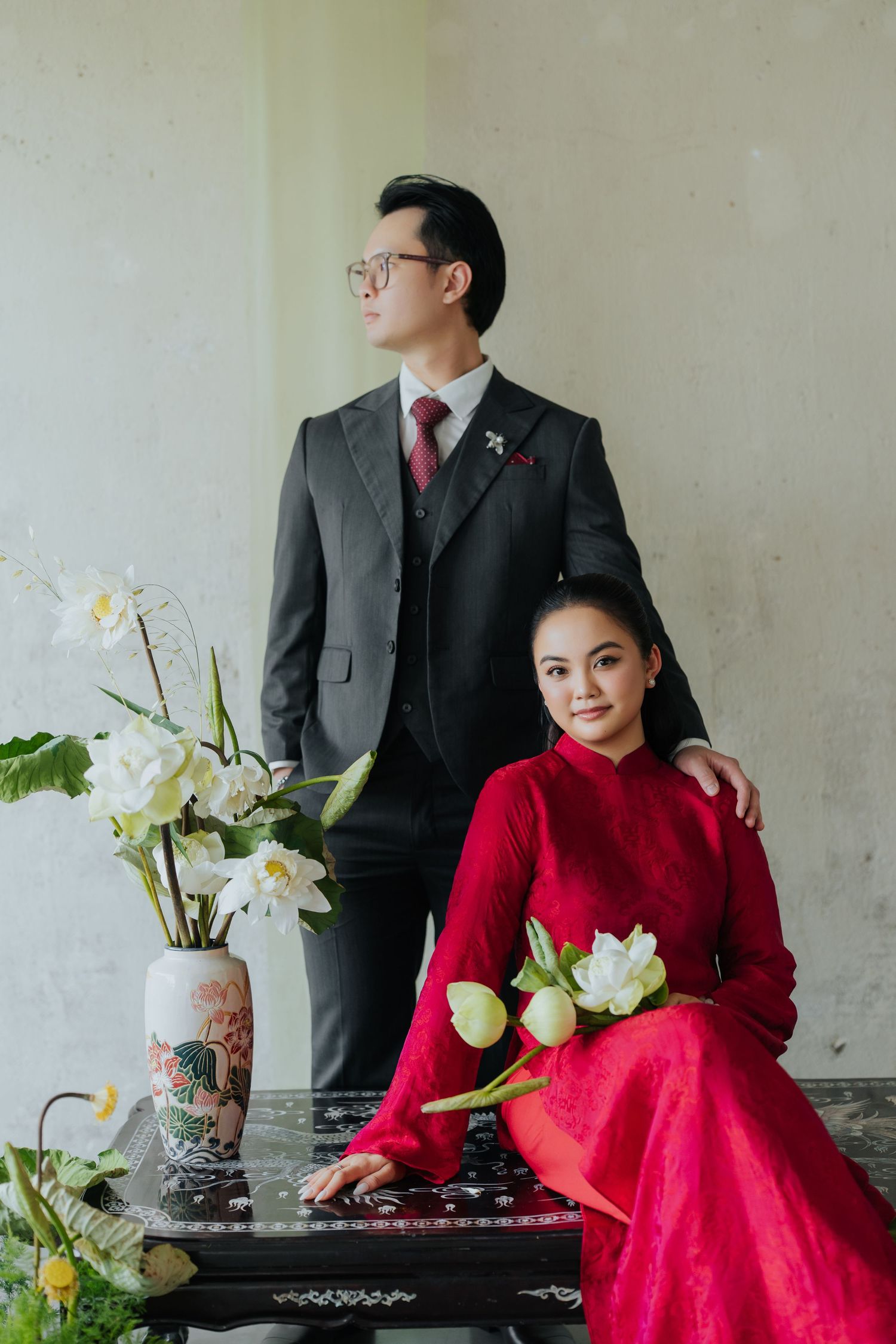 A couple in traditional Vietnamese wedding attire, man in dark suit, woman in red ao dai holding white lotus flowers.