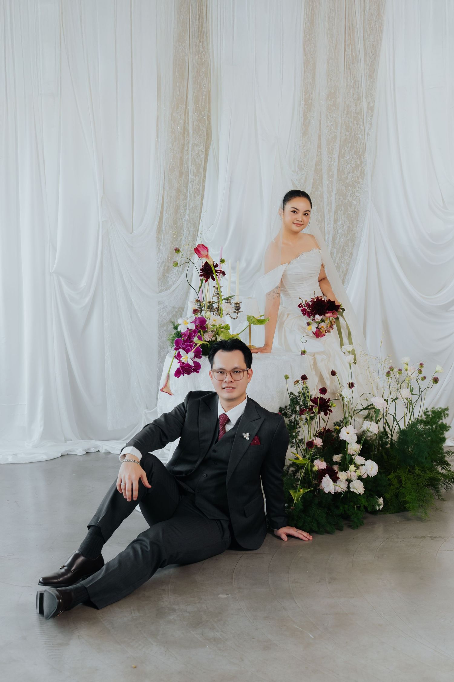 A groom in a black suit sits on the floor while his bride in a white gown stands behind floral arrangements.