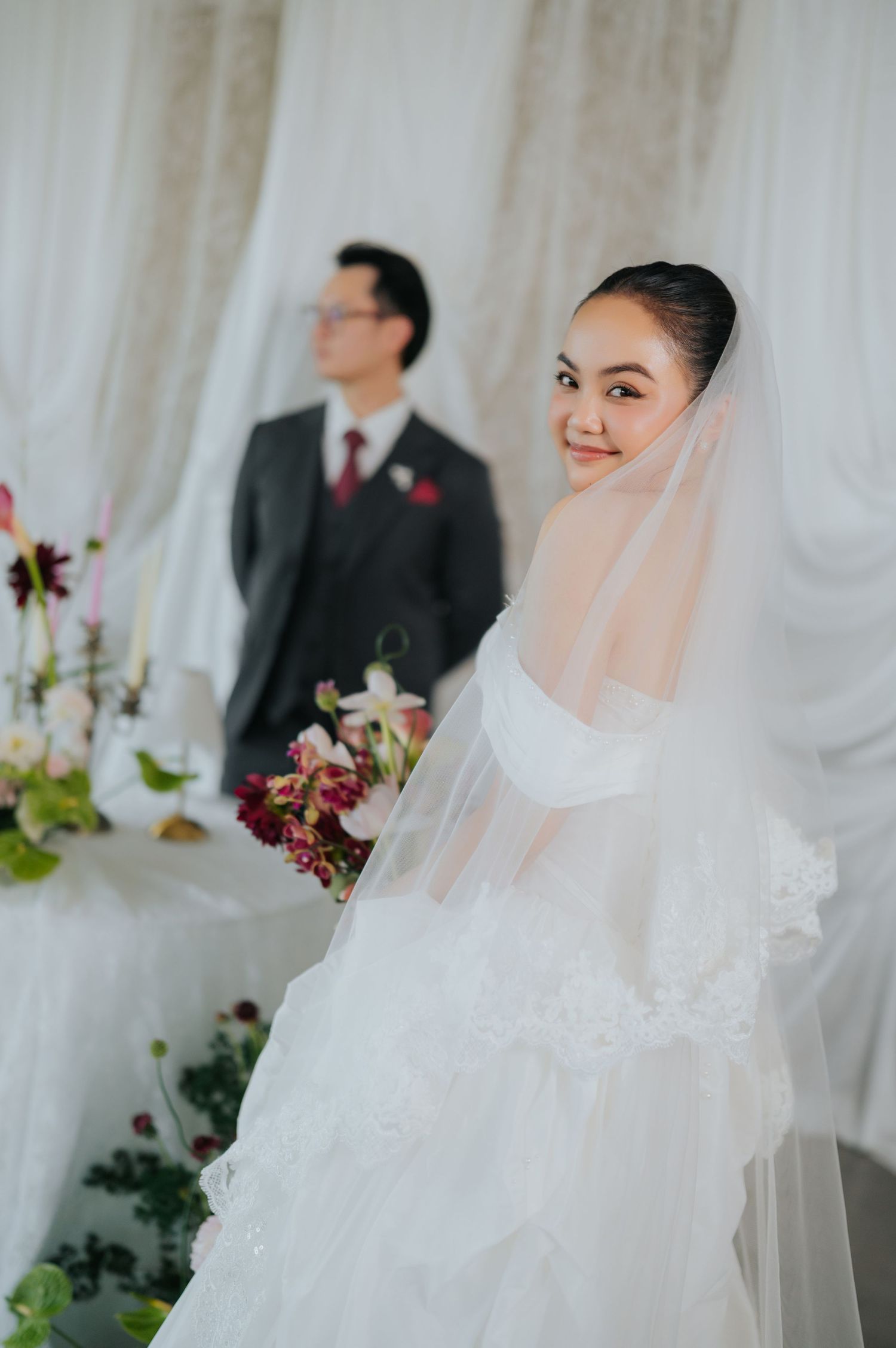 A smiling bride in a white gown and veil holds a bouquet while a groom in a gray suit stands in the background.