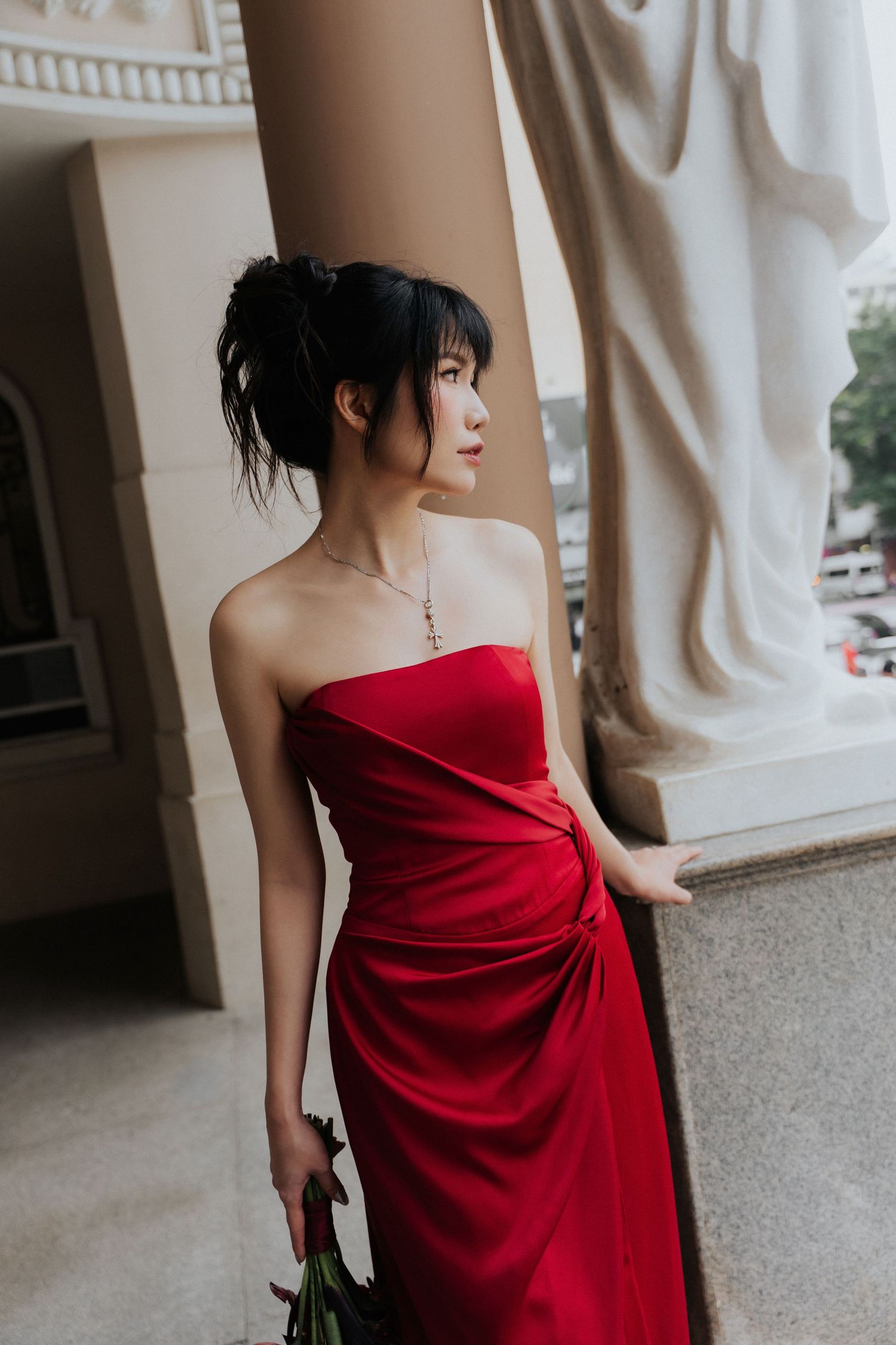Young woman in red strapless satin gown holding roses, posing by ornate column in elegant building.