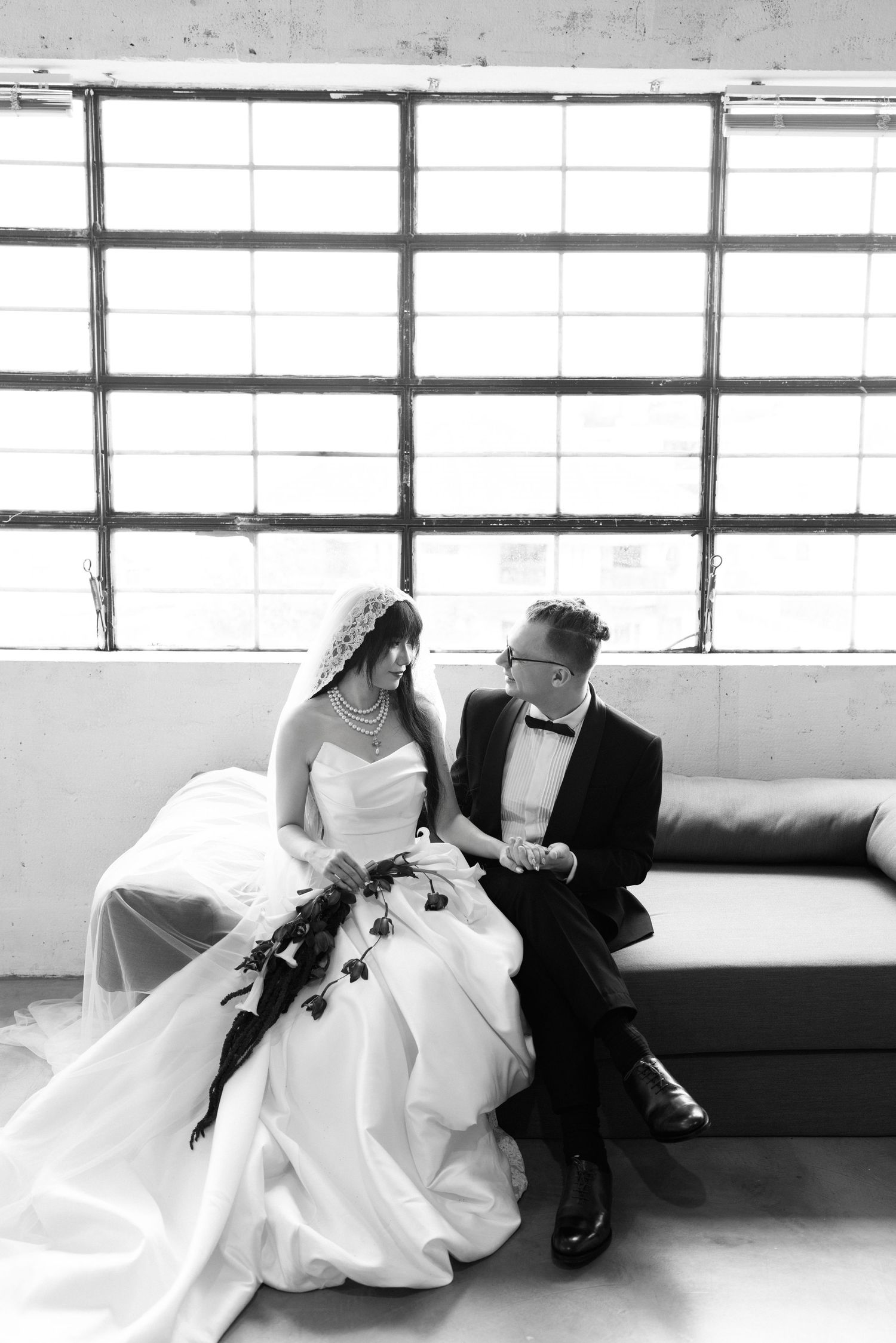 Black and white photo of bride and groom sitting together on a couch, sharing a loving glance in front of large windows.