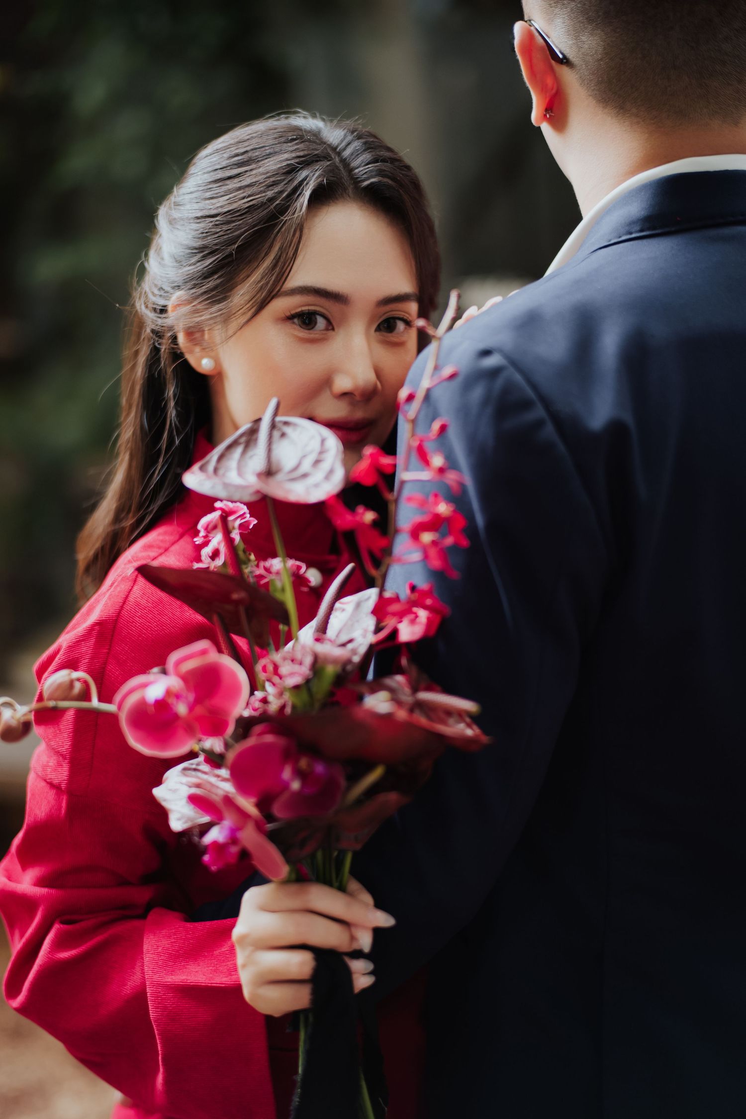 A woman in a red dress holds pink and red flowers, gazing at a man in a navy suit during a romantic outdoor moment.