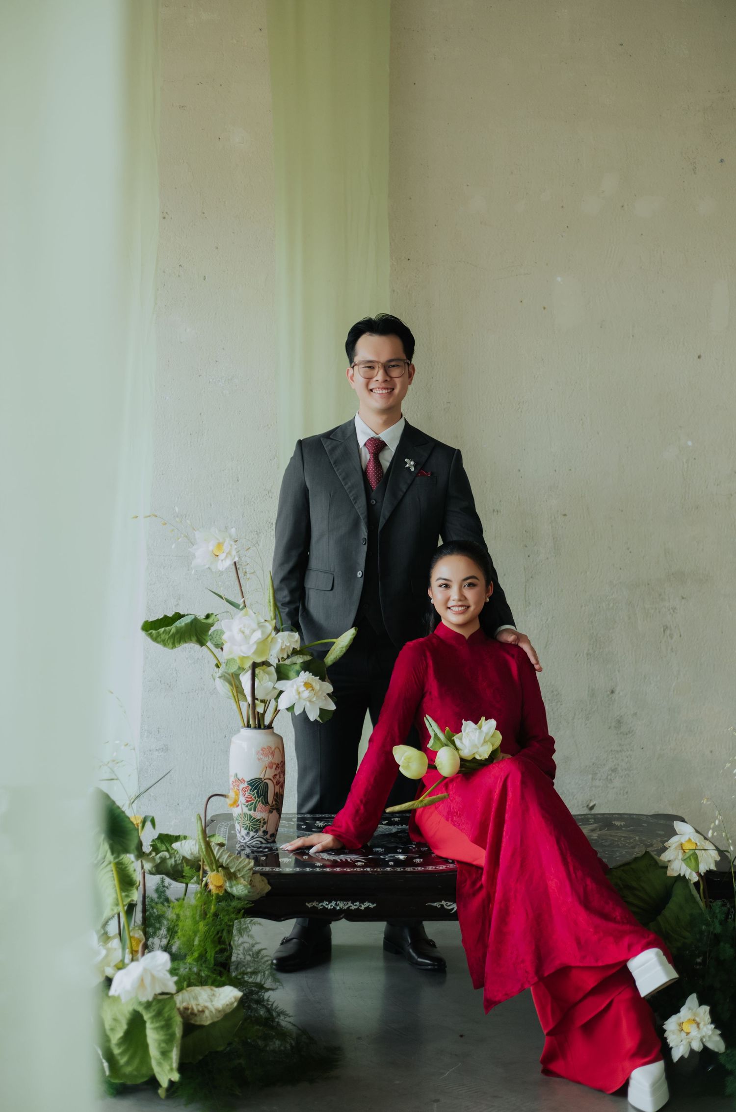 Couple in Vietnamese traditional ao dai and suit posing with white flowers and ornate vase in elegant studio setting.