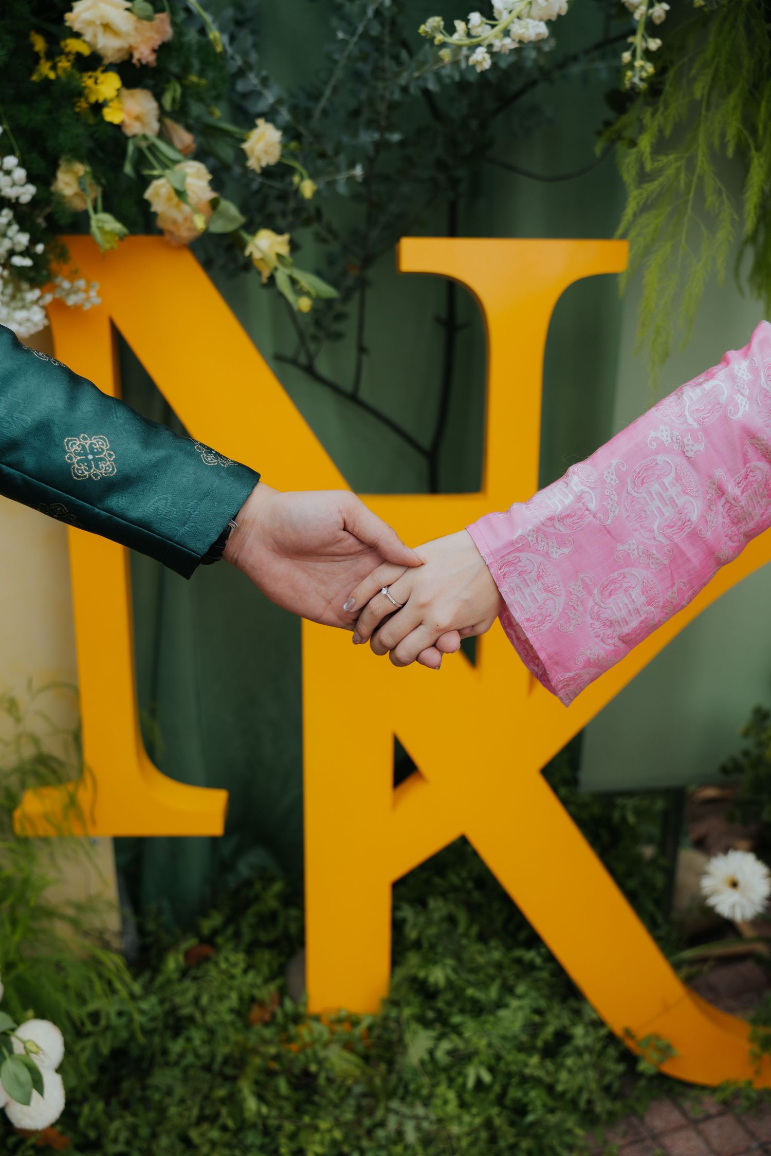 Couple holding hands in traditional attire before yellow monogram letters N and K, surrounded by floral decor.