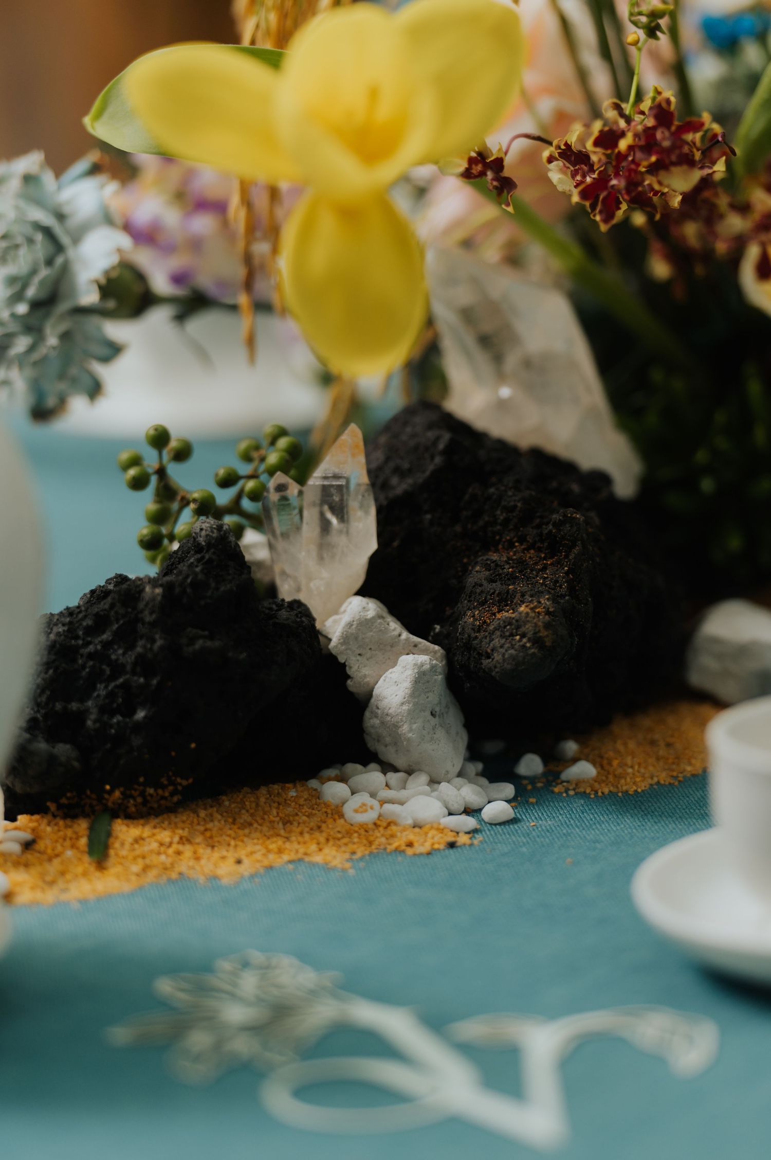 Crystal quartz and lava rock centerpiece with yellow orchids on teal tablecloth for elegant event decor.